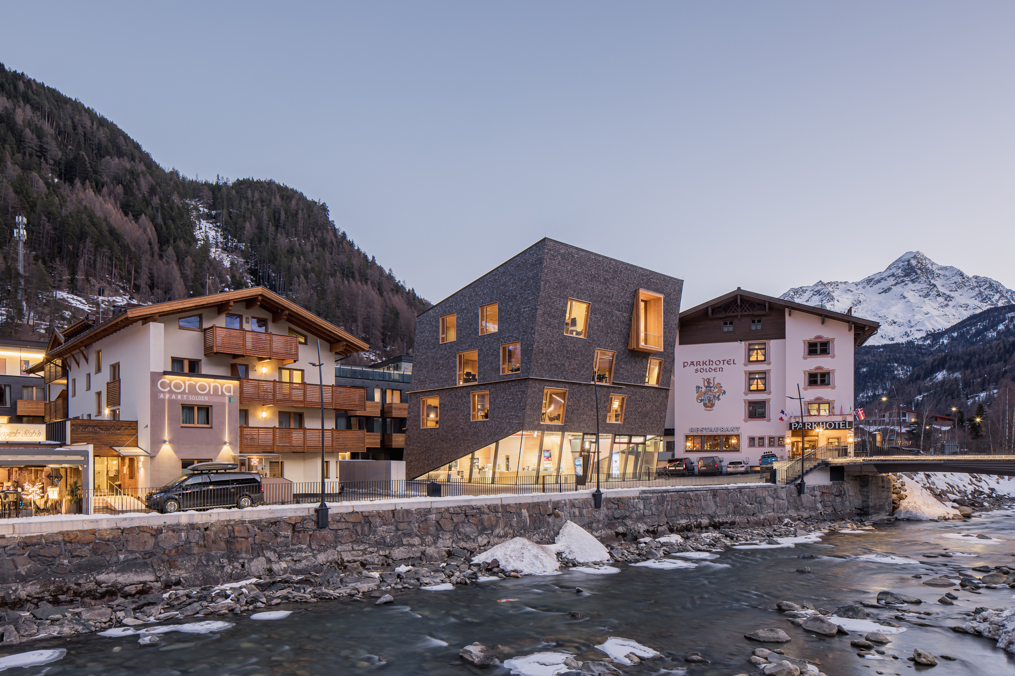 Cubic volume clad in dark shingles nestled between traditional alpine buildings along a winter stream