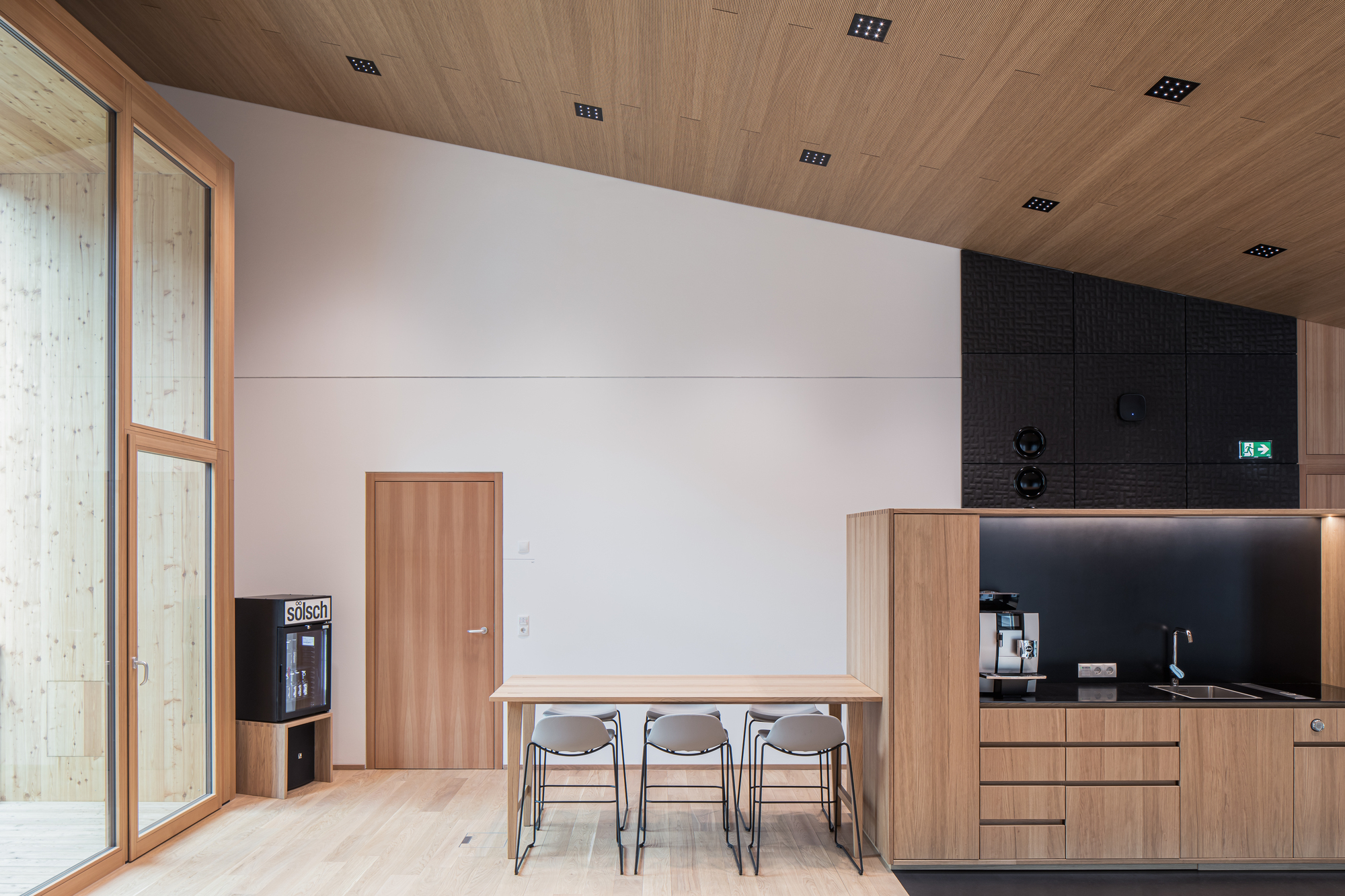 Timber dining table with grey chairs beside wood door and coffee bar under slatted ceiling