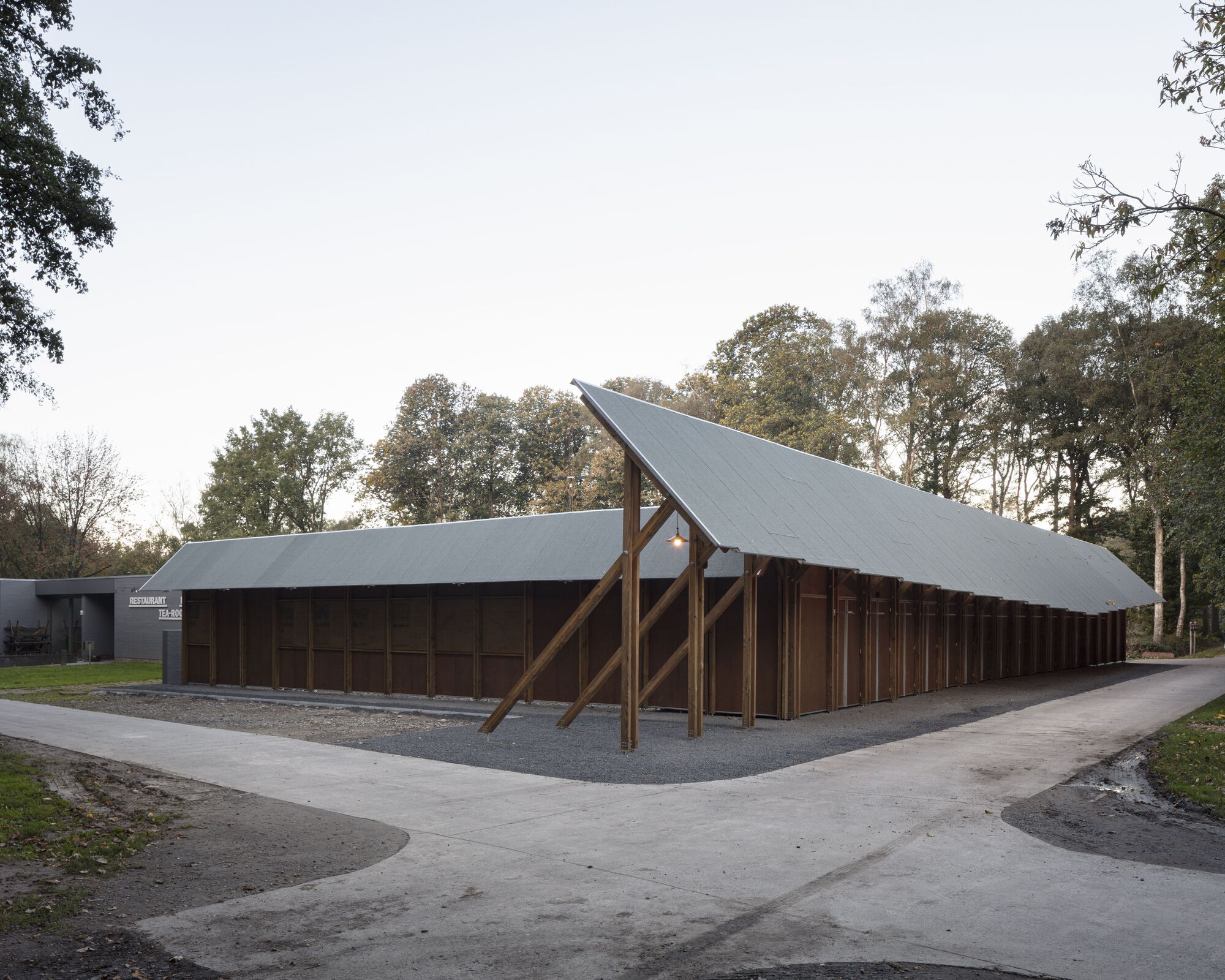 Long pavilion with timber portal frames and metal roofing beside a paved path on an overcast autumn day