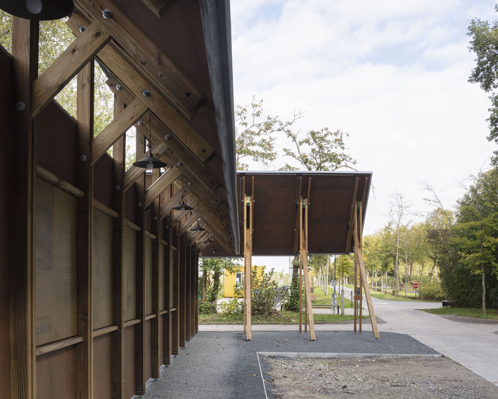 Timber canopy structure with slanted columns supporting a raised dark wood volume on stilts
