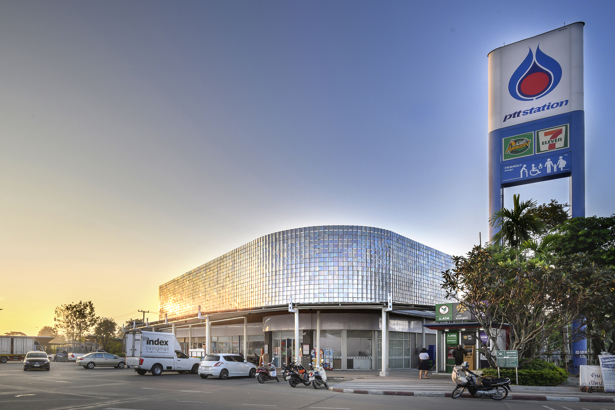 Rounded translucent glass panel facade above a canopy with vehicles and motorcycles at dusk