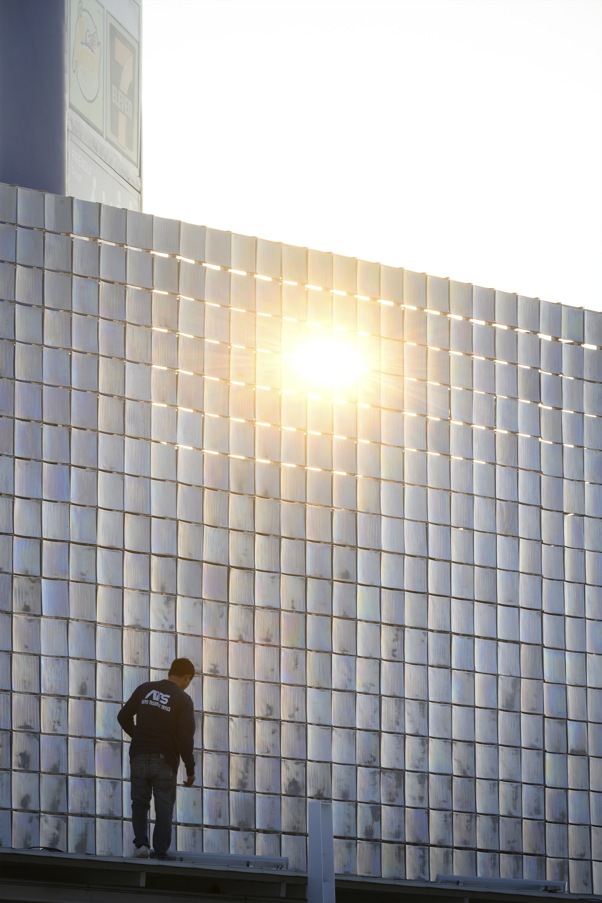 Figure standing on a rooftop platform beside the backlit glass tile facade with sun flare