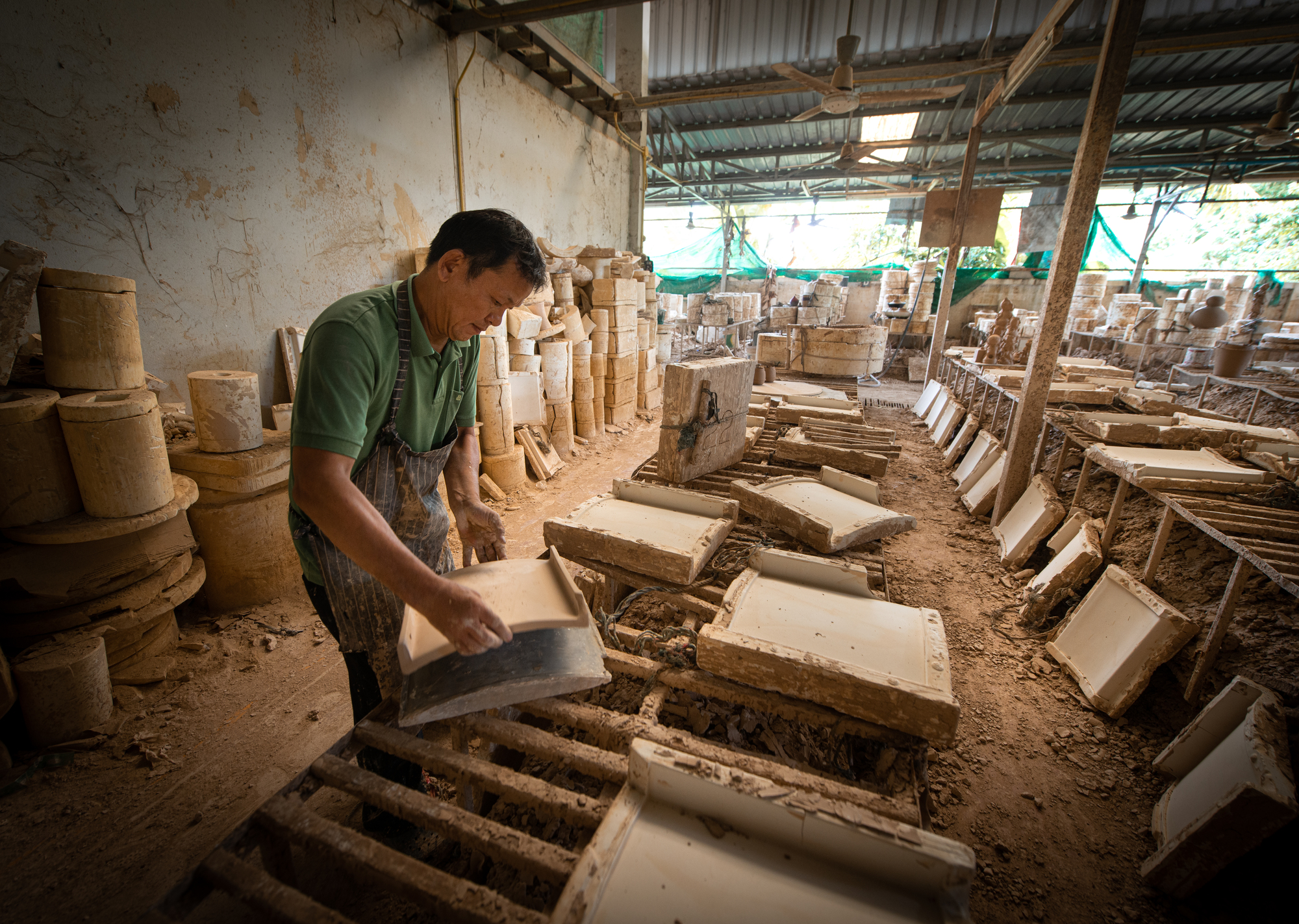 Worker placing ceramic tiles onto wooden drying racks beneath a corrugated metal roof