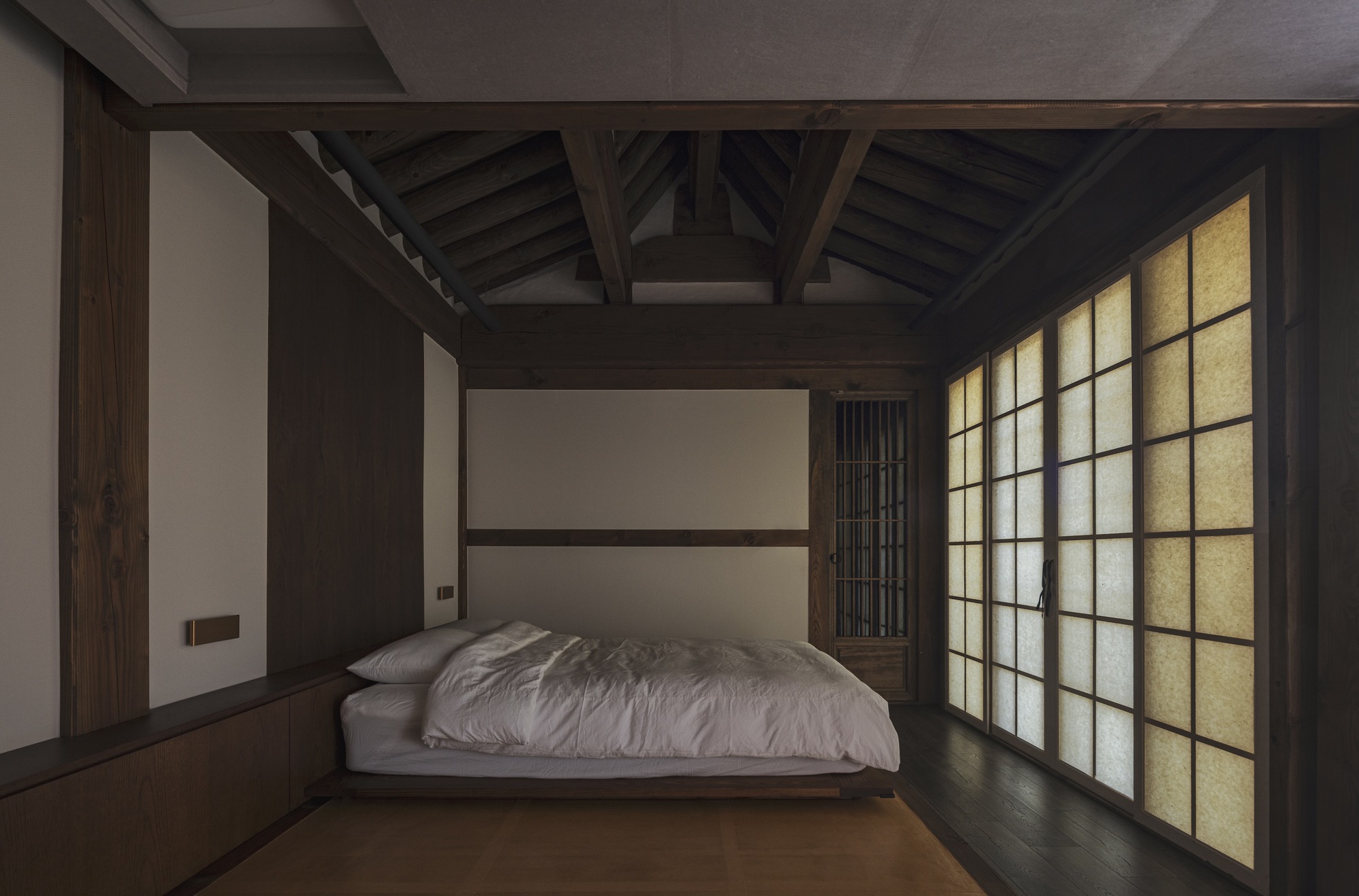 Bedroom with exposed roof trusses and backlit paper screens casting soft light at dusk