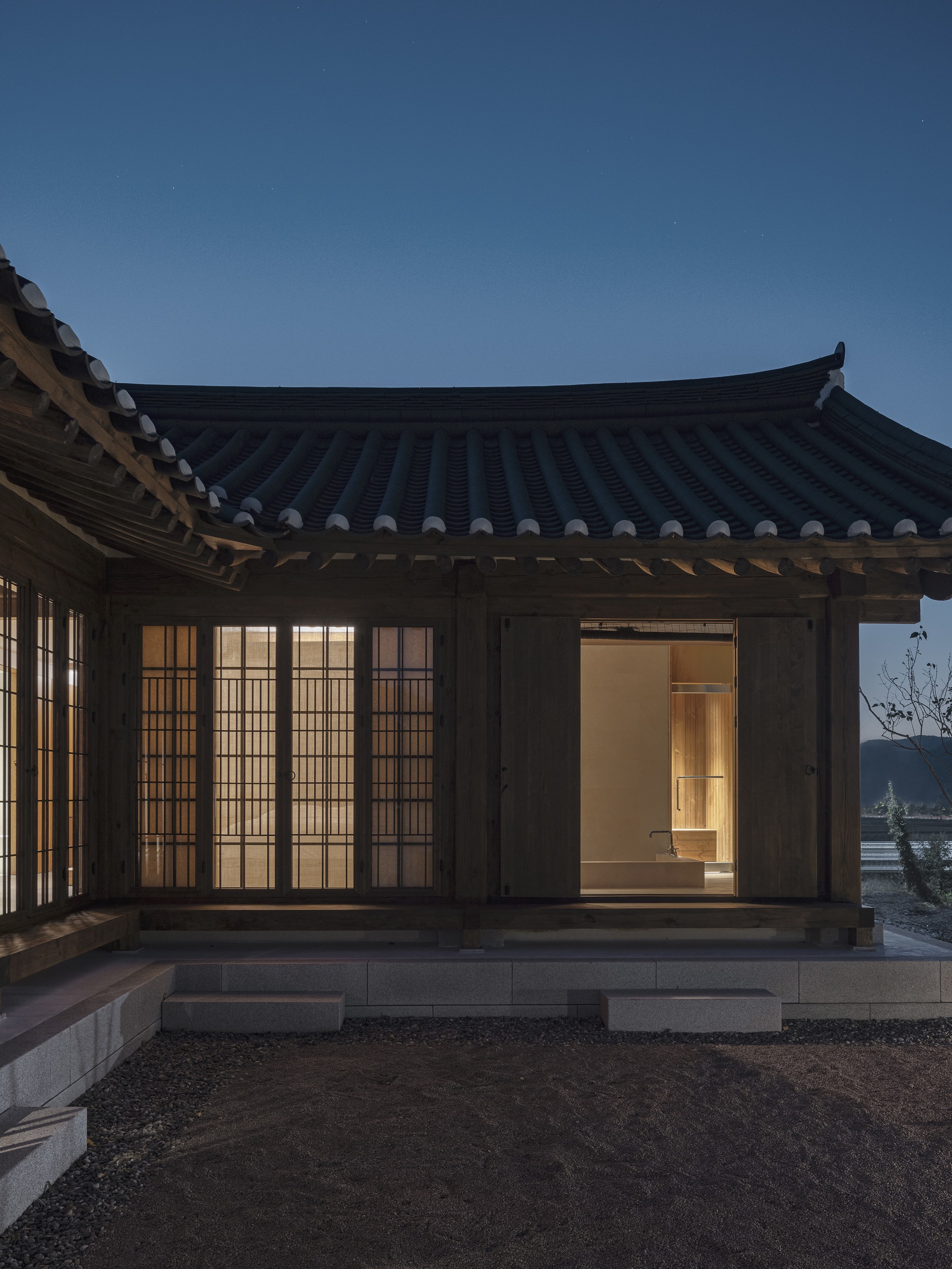 Timber pavilion with curved tiled roof and translucent gridded screens illuminated at dusk