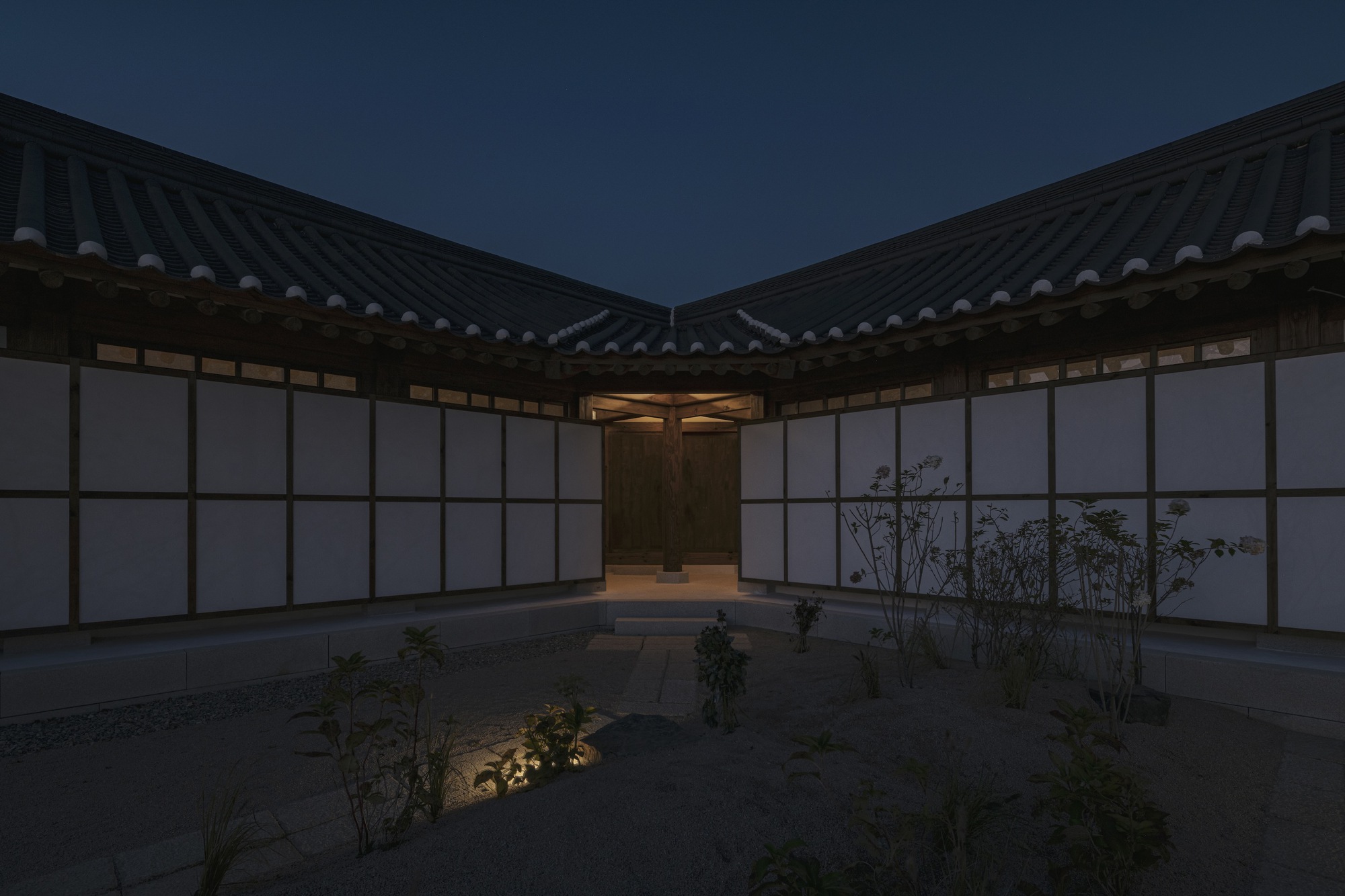 Interior courtyard with translucent wall panels and planted beds beneath converging tiled rooflines at twilight