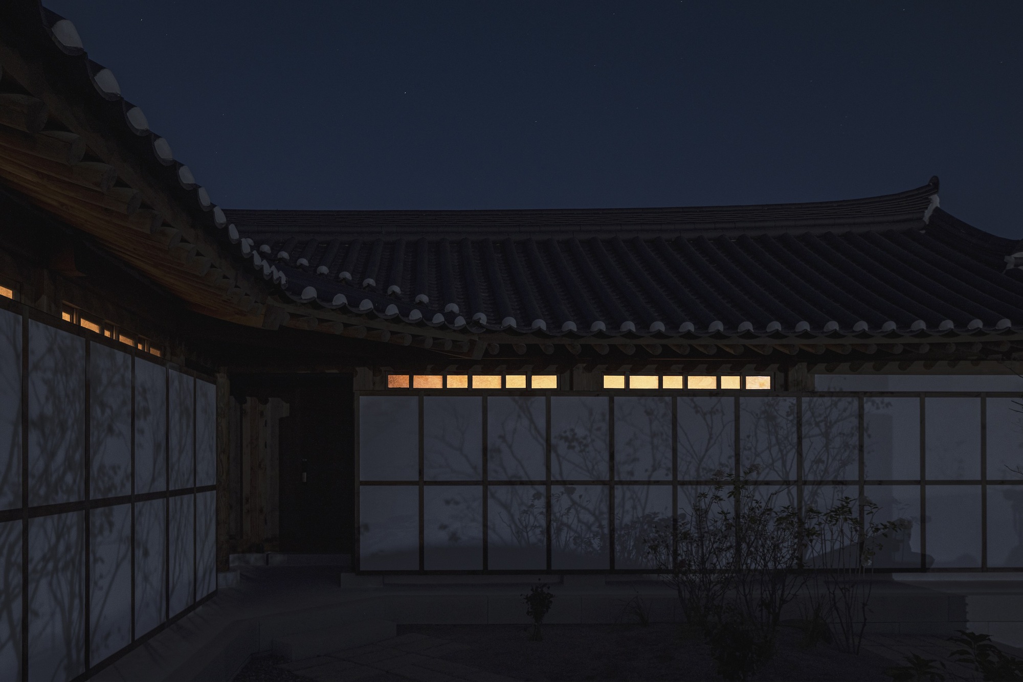 Courtyard facade with translucent panels and illuminated clerestory windows under sweeping tiled eaves at dusk