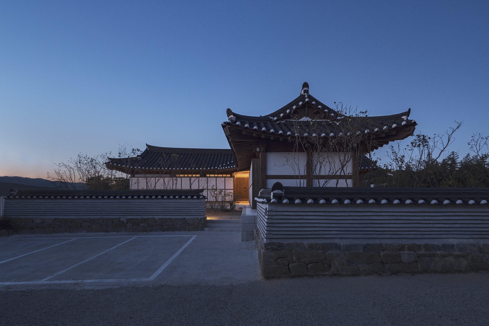 Entry gate and timber pavilions with curved tiled roofs framed by bare winter trees at sunset