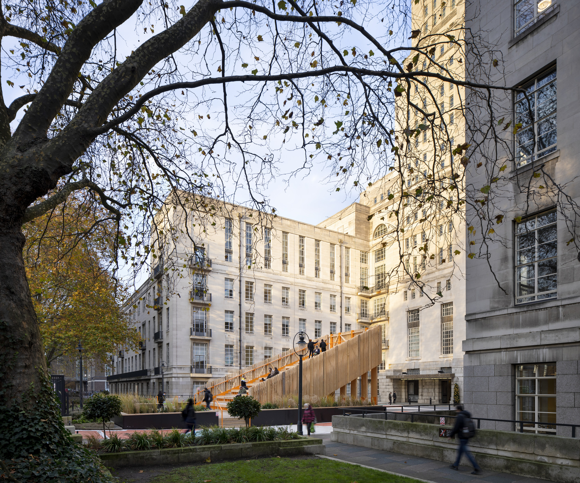 Timber pedestrian bridge crossing a courtyard garden framed by bare tree branches in autumn