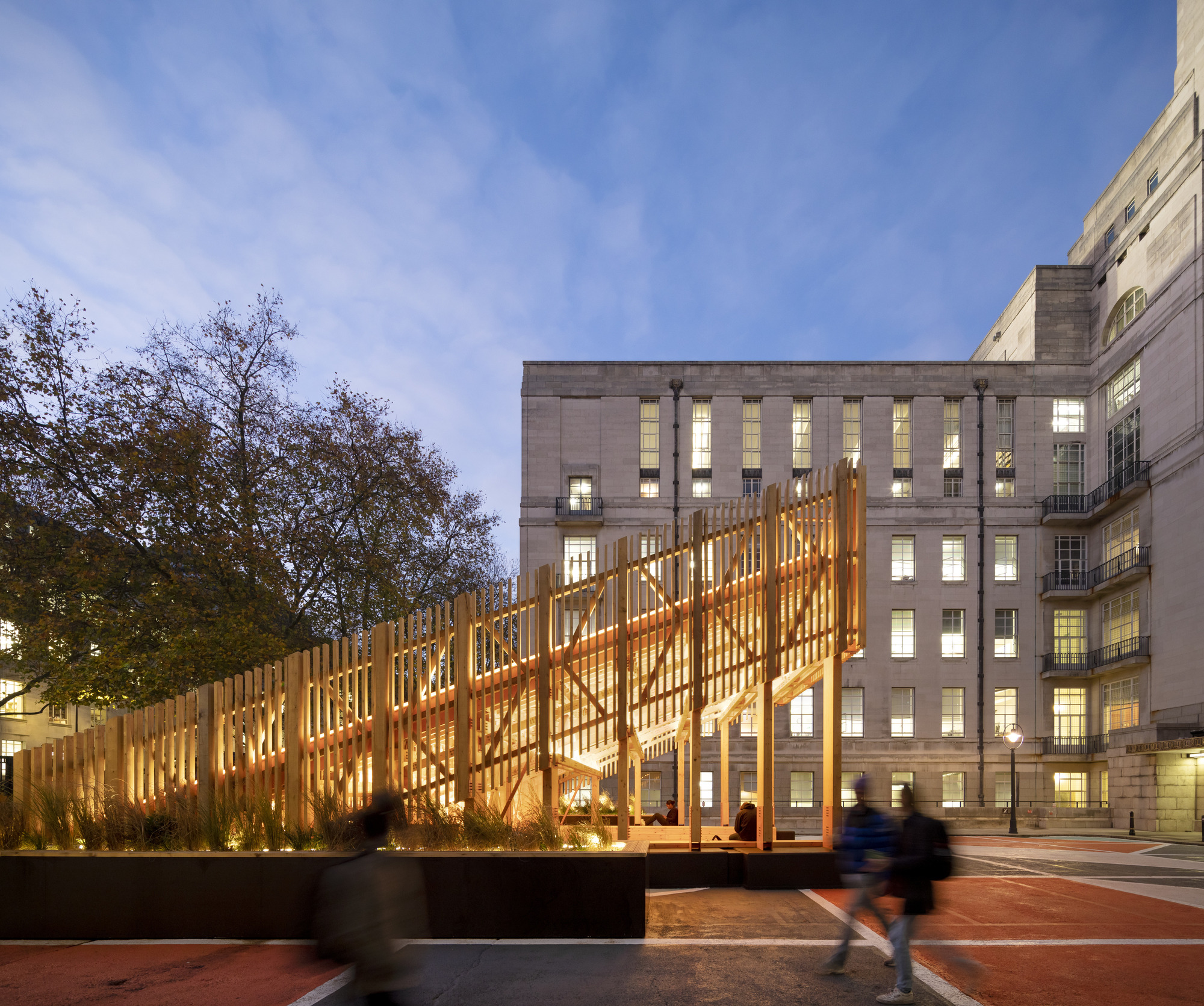 Long view of the illuminated timber framework extending across the courtyard at dusk