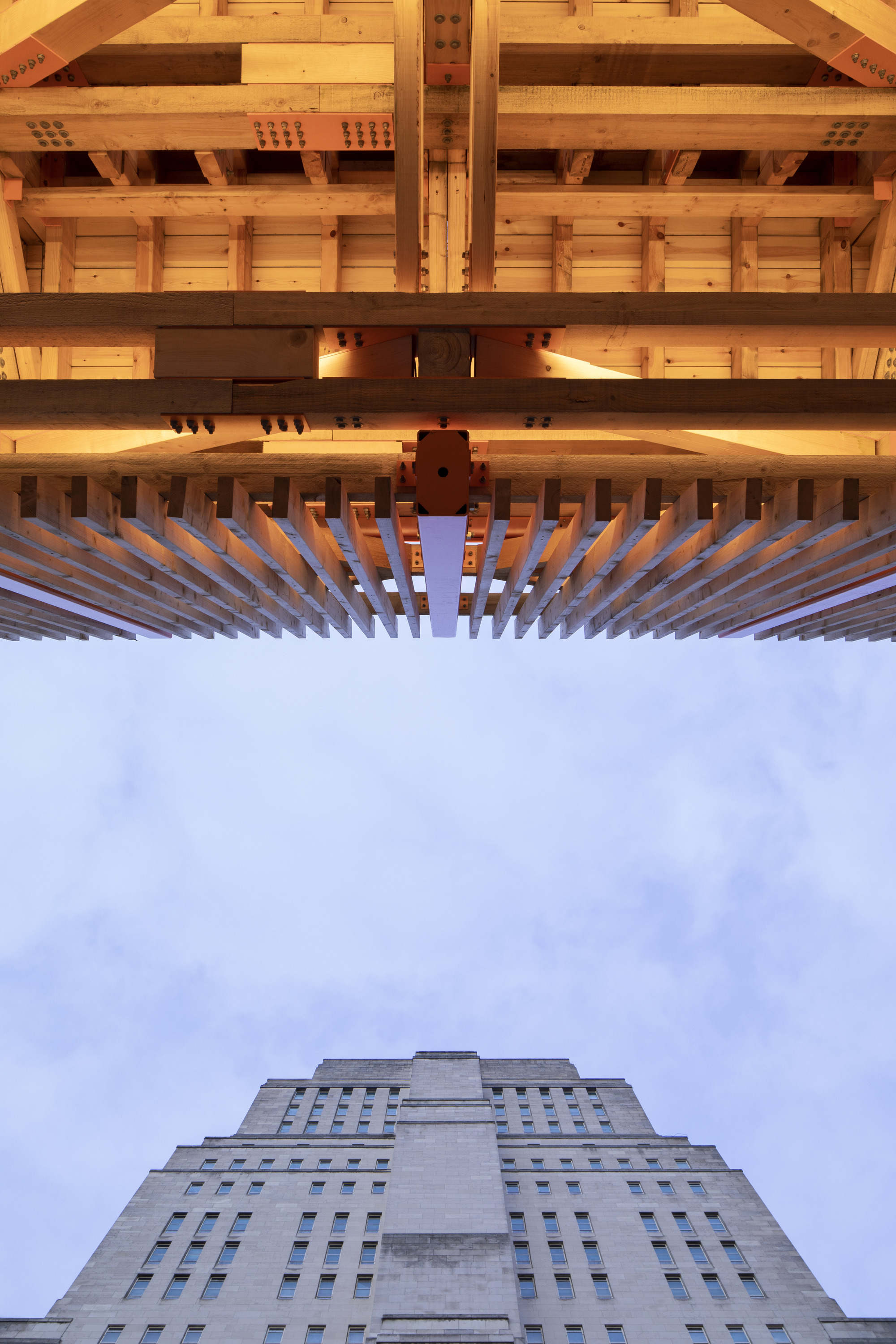 Upward view through layered timber joists and beams with a stepped concrete tower beyond