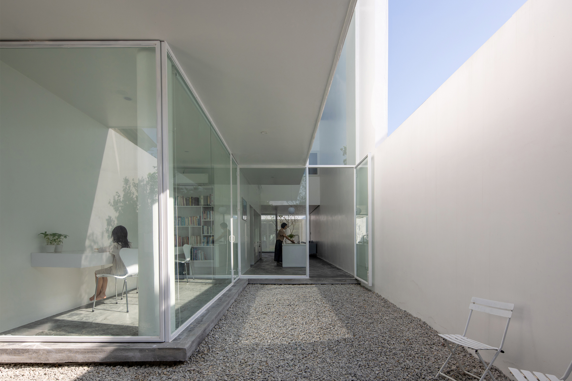 Narrow gravel courtyard framed by glass walls with a figure inside and blue sky above