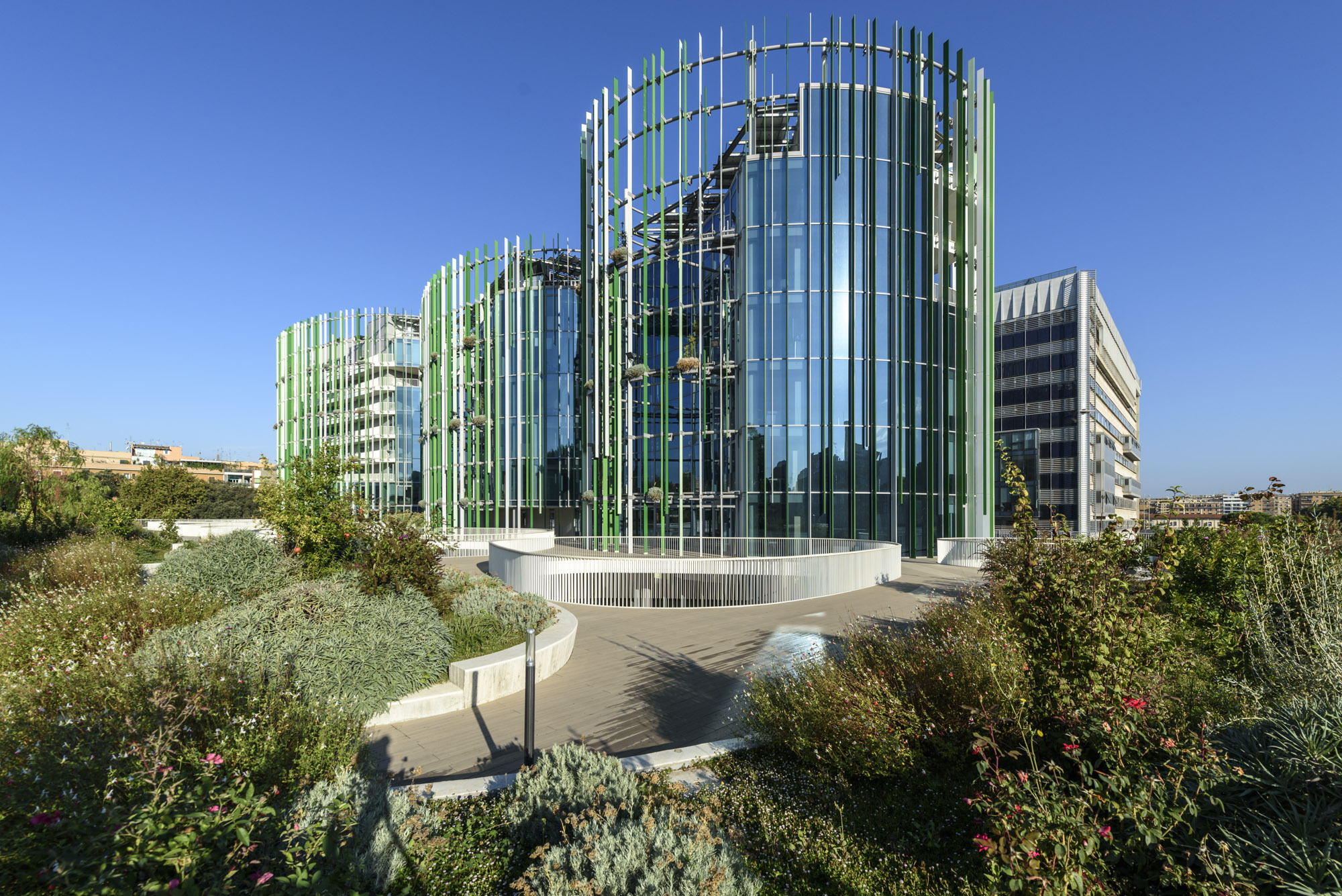 Glass tower wrapped in vertical green and white fins rising from a landscaped garden terrace under clear skies