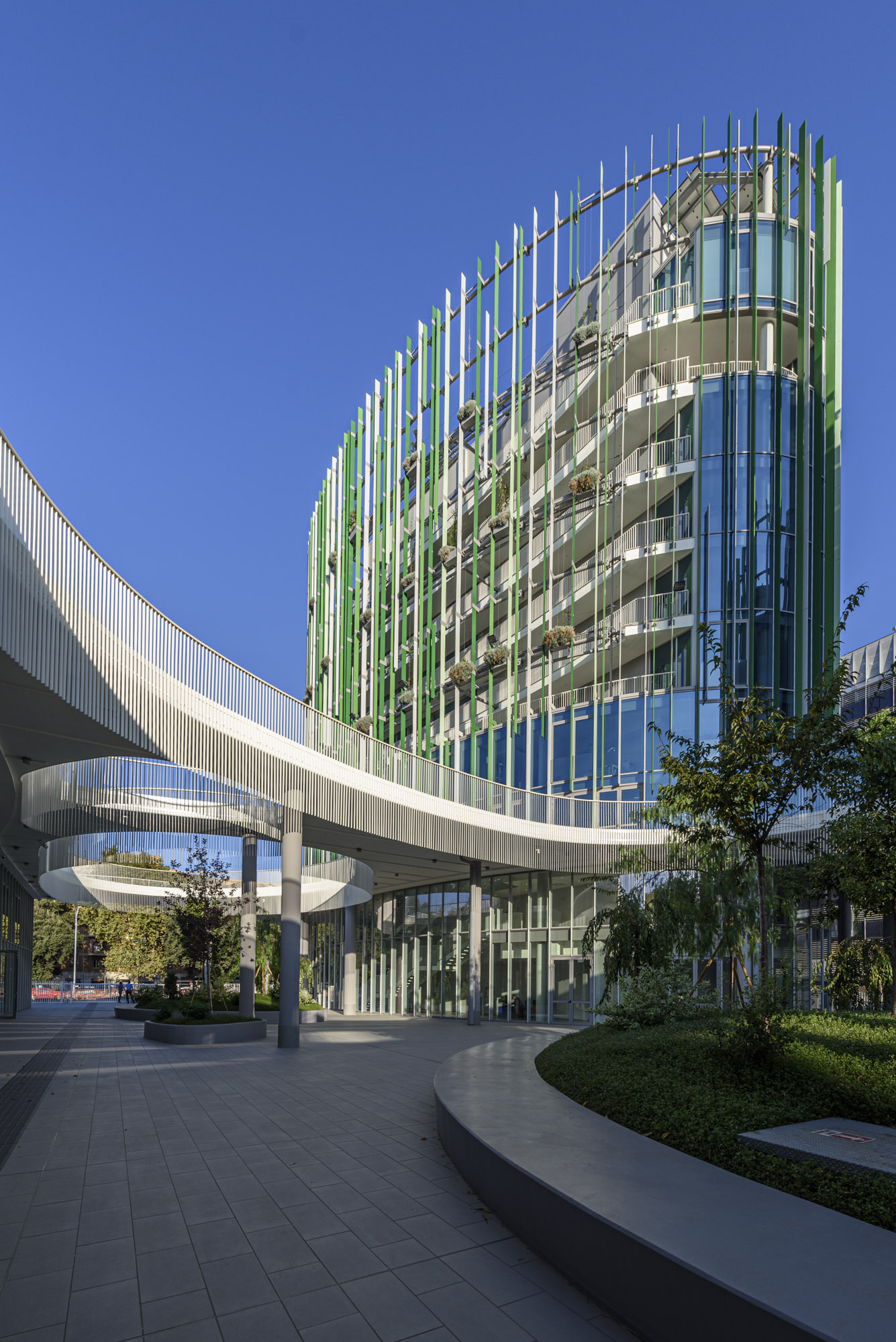 Ground-level view of the glazed tower with vertical fin screen and elevated walkway spanning above planted beds