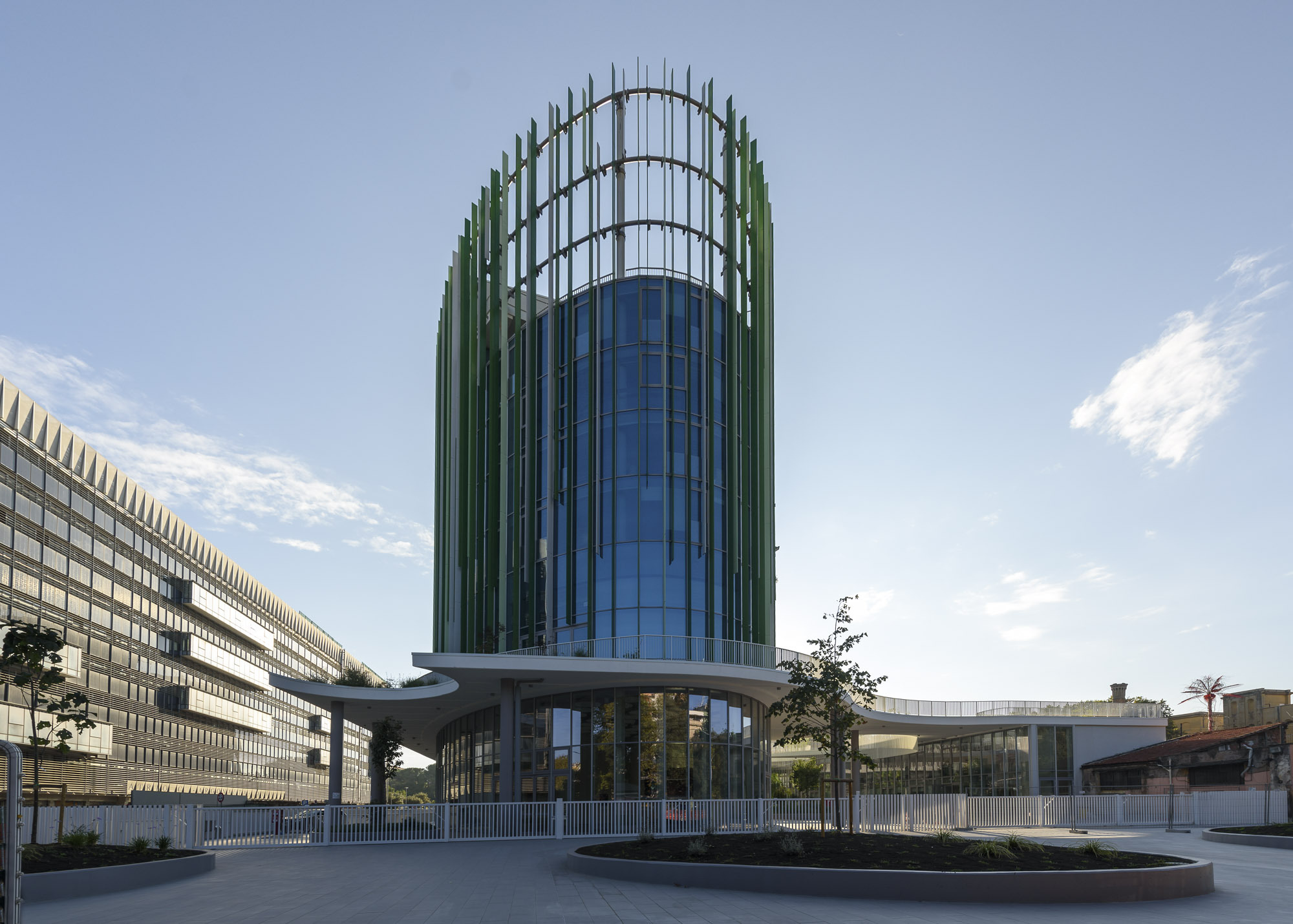 Cylindrical glass tower with vertical fin cladding viewed from the plaza with elevated pedestrian bridge at base
