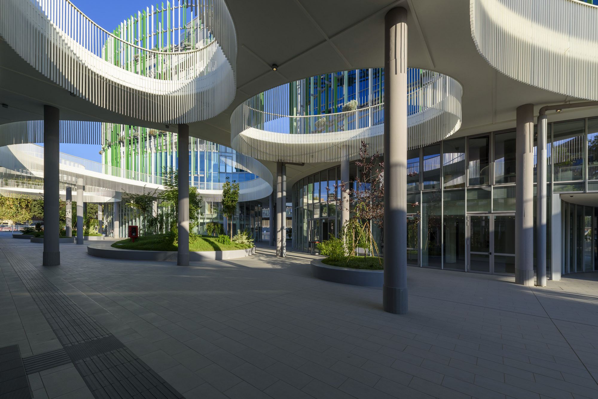 Covered entrance plaza with cylindrical columns, curved canopy and planted beds beneath a blue sky