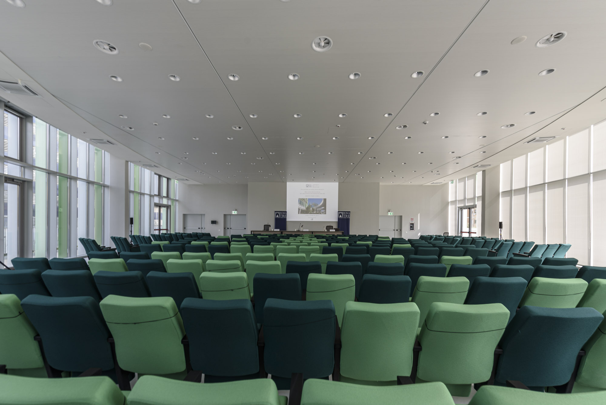Auditorium interior with rows of green upholstered seating beneath white ceiling panels and recessed lighting