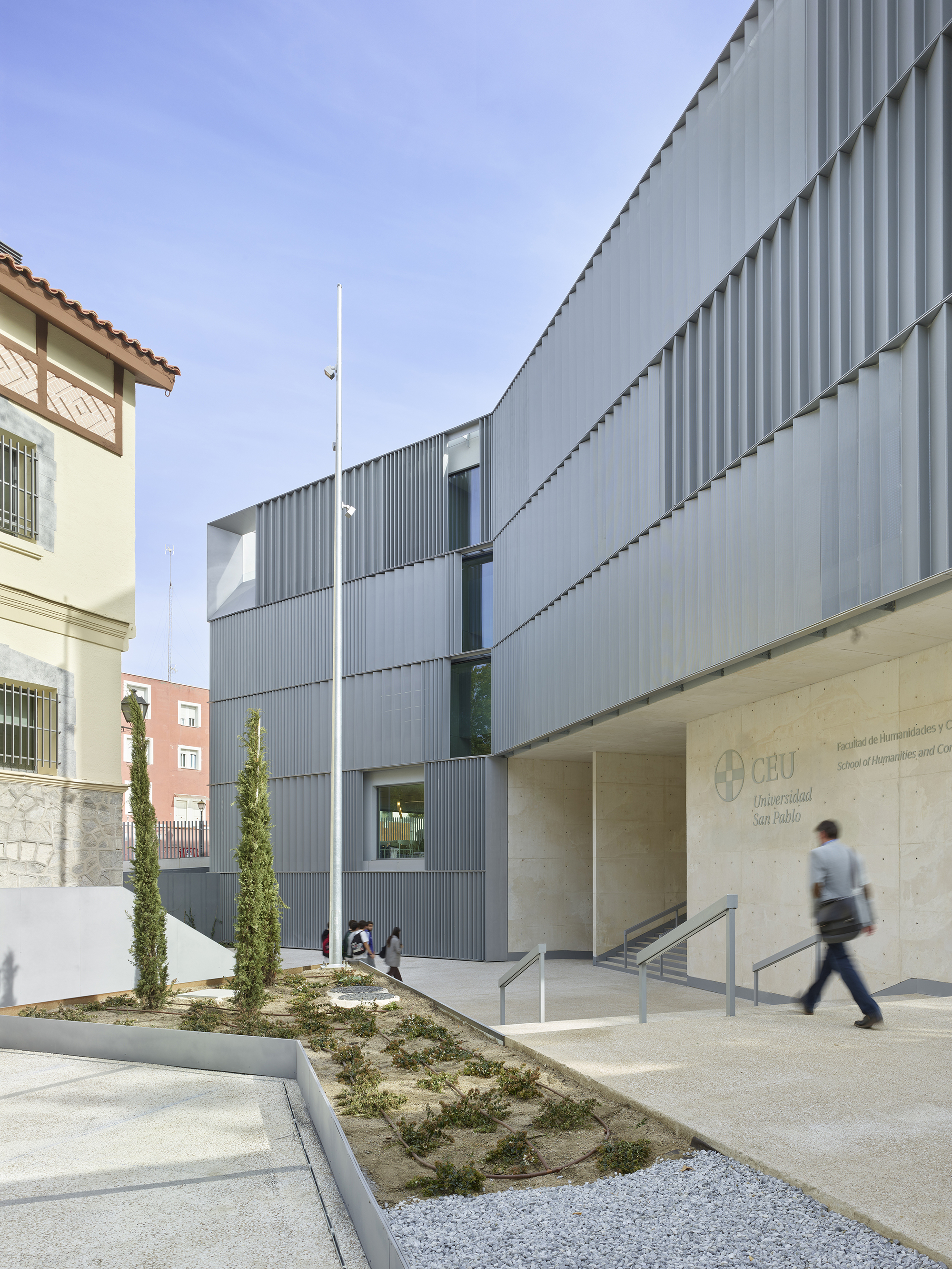Entrance courtyard with corrugated metal facade, planted beds with cypresses, and a person walking past