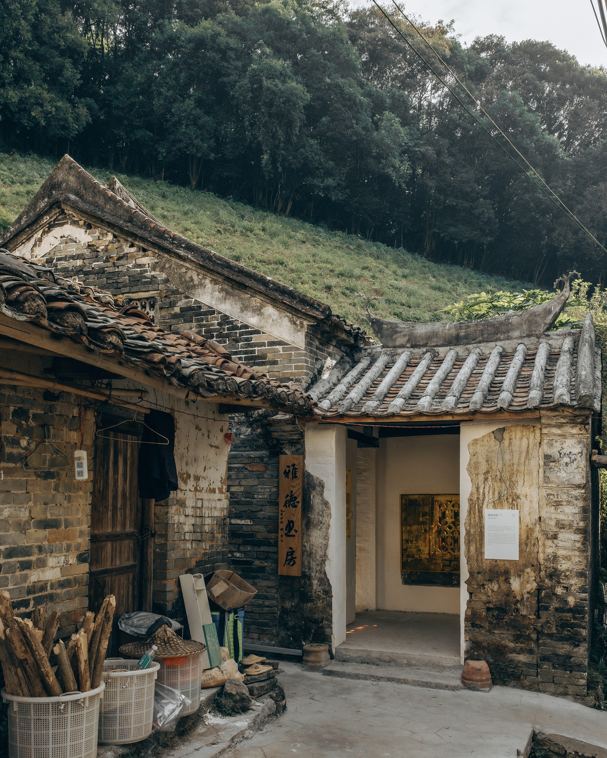 Weathered brick courtyard entrance with curved tile roofs and stacked firewood beneath a forested hillside