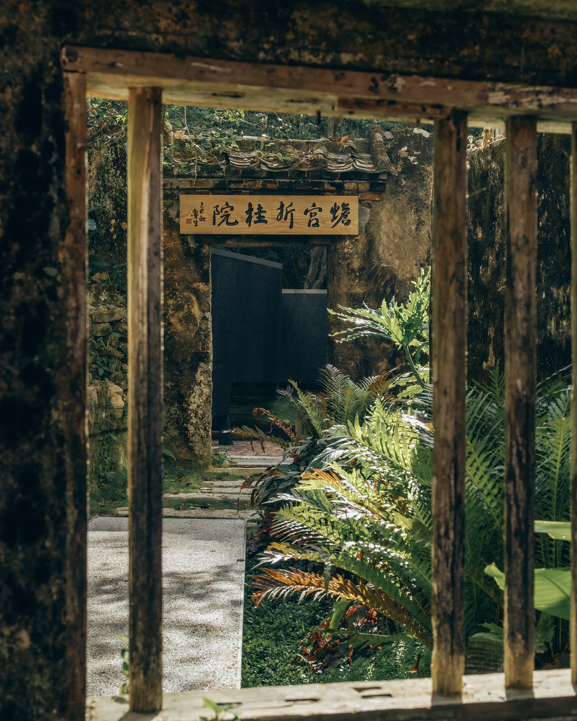 Timber colonnade framing a courtyard path with carved wooden signage and tropical ferns in dappled sunlight