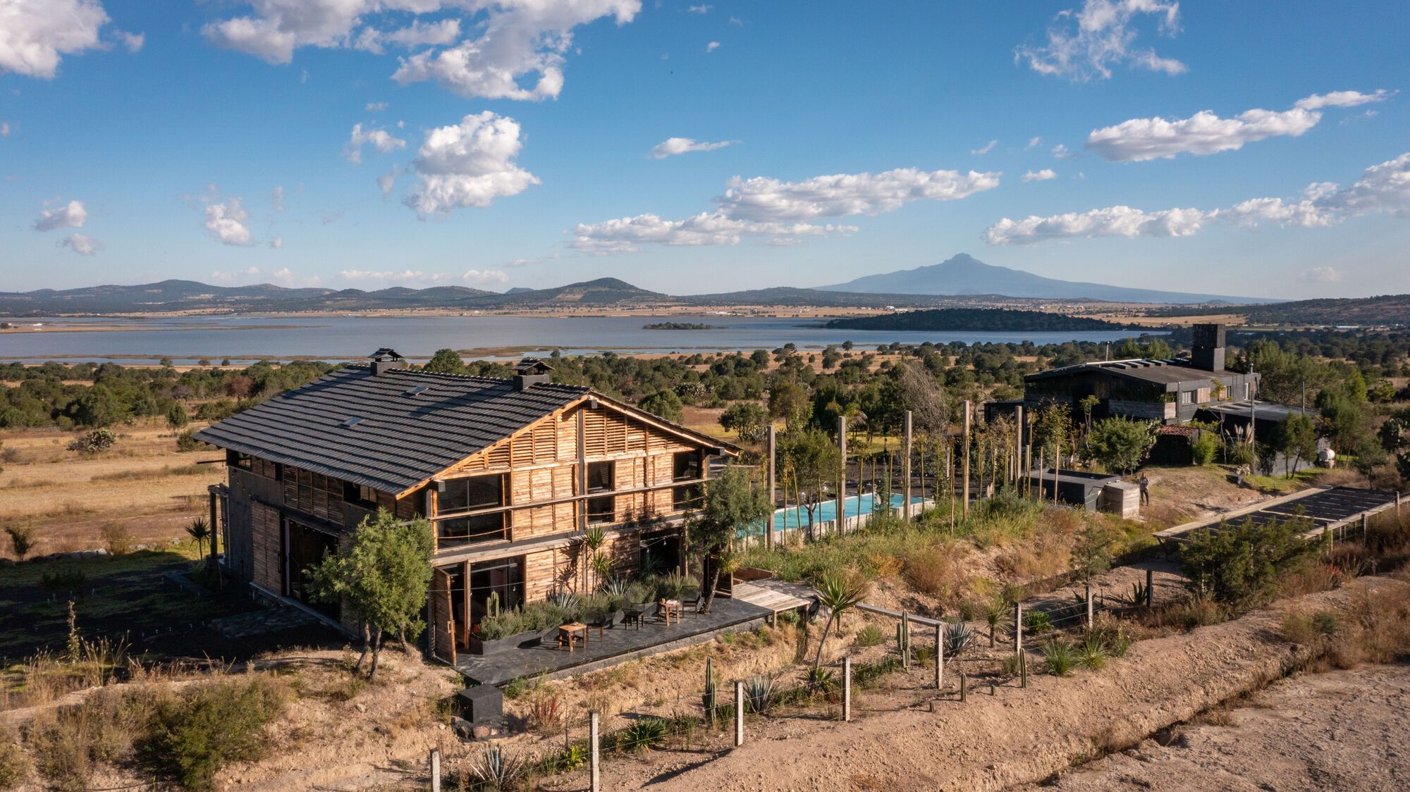 Aerial view of the timber-clad two-storey house with corrugated metal roof overlooking a distant lake and mountains
