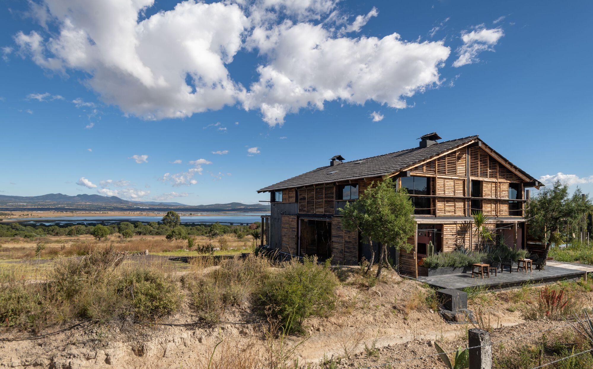 Timber slatted facade with gable roof overlooking native grassland and distant lake under white clouds