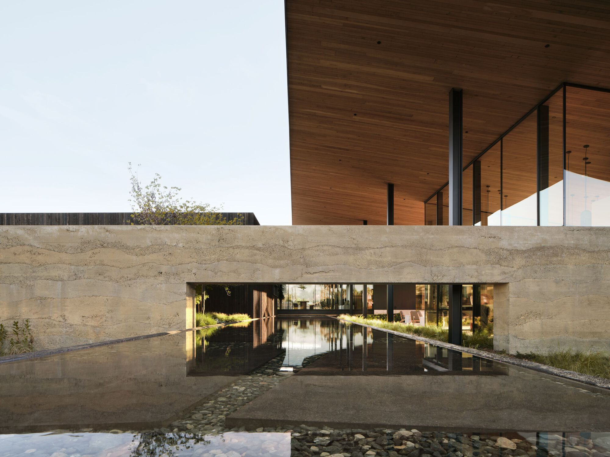 Rammed earth wall bridging a reflecting pool beneath timber soffit with steel columns at twilight