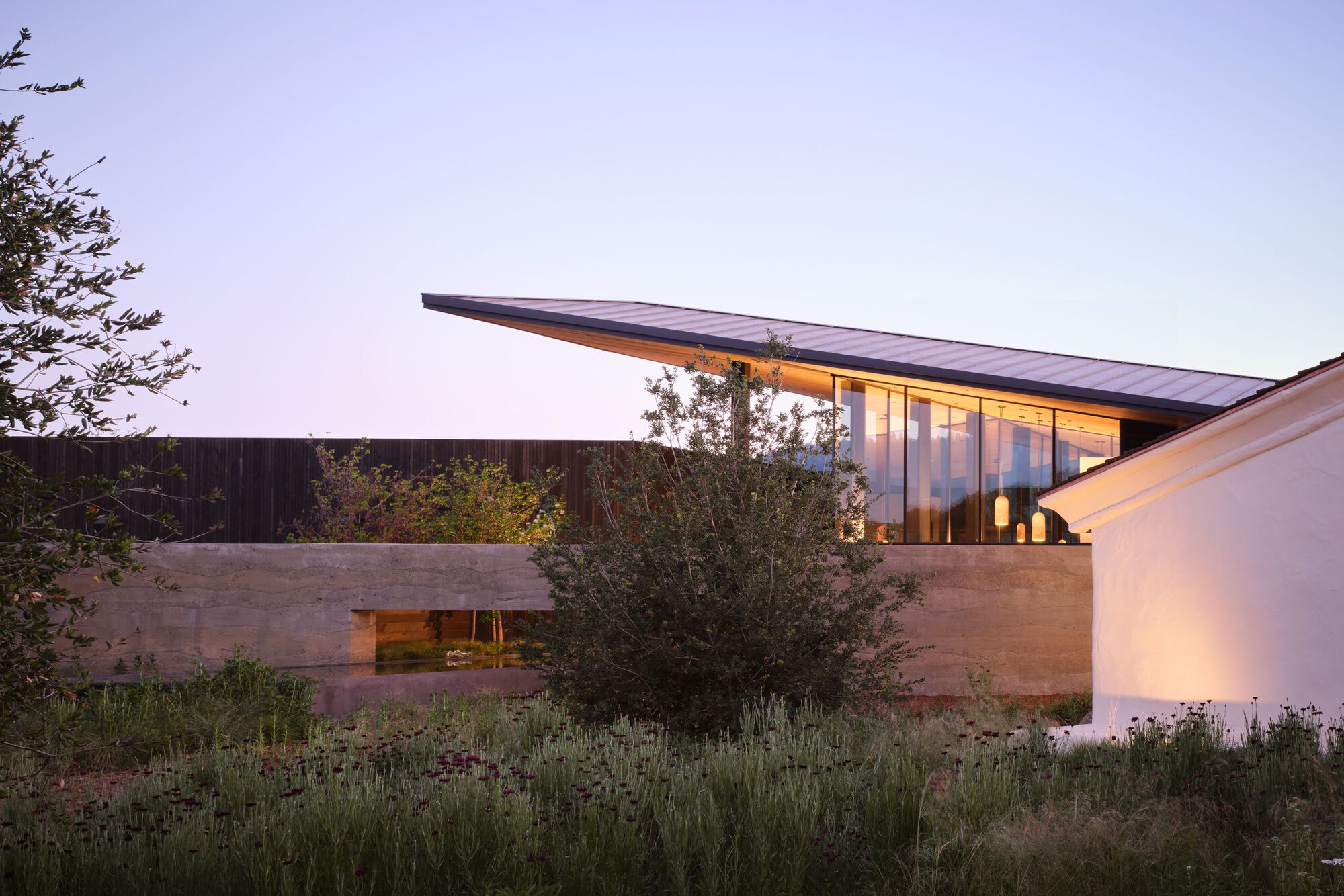 Exterior view of the cantilevered roof and rammed earth walls among drought-tolerant plantings at dusk