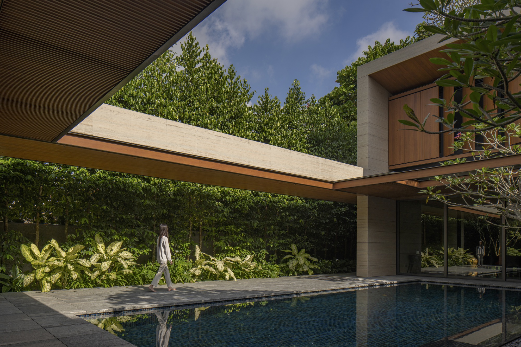 Pool terrace with a figure walking past dense plantings beneath cantilevered concrete and timber volumes