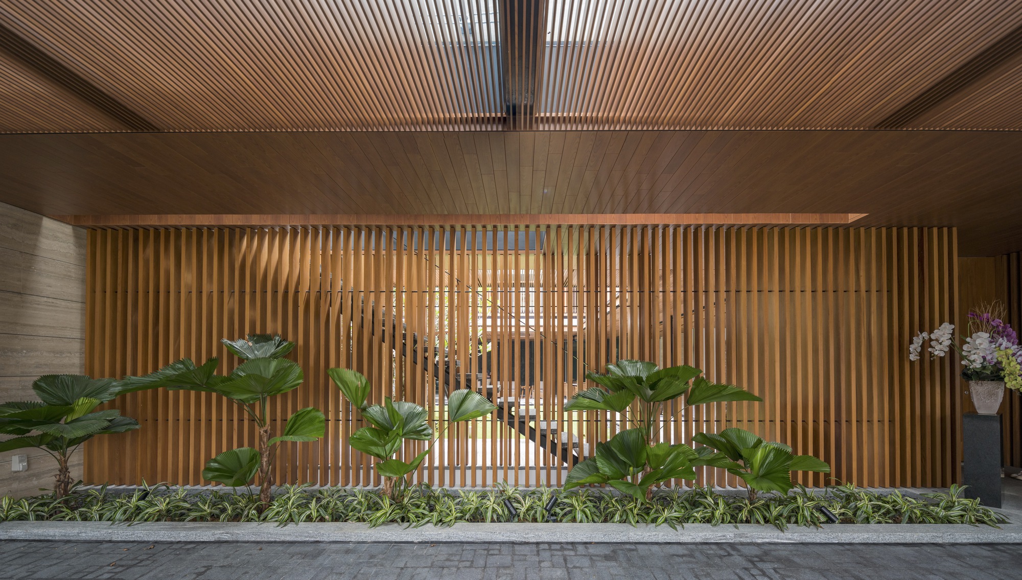 Entry facade with vertical timber screen and planted bed of broad-leafed palms at dusk