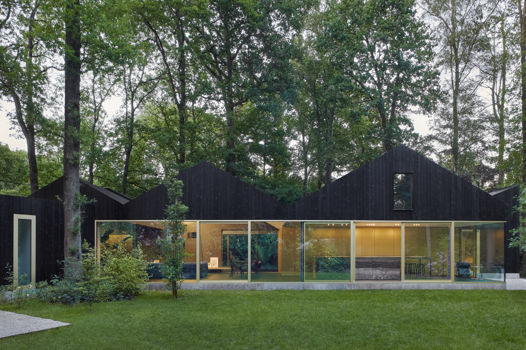 Garden facade with continuous glazing beneath charred timber volumes and gabled rooflines at dusk