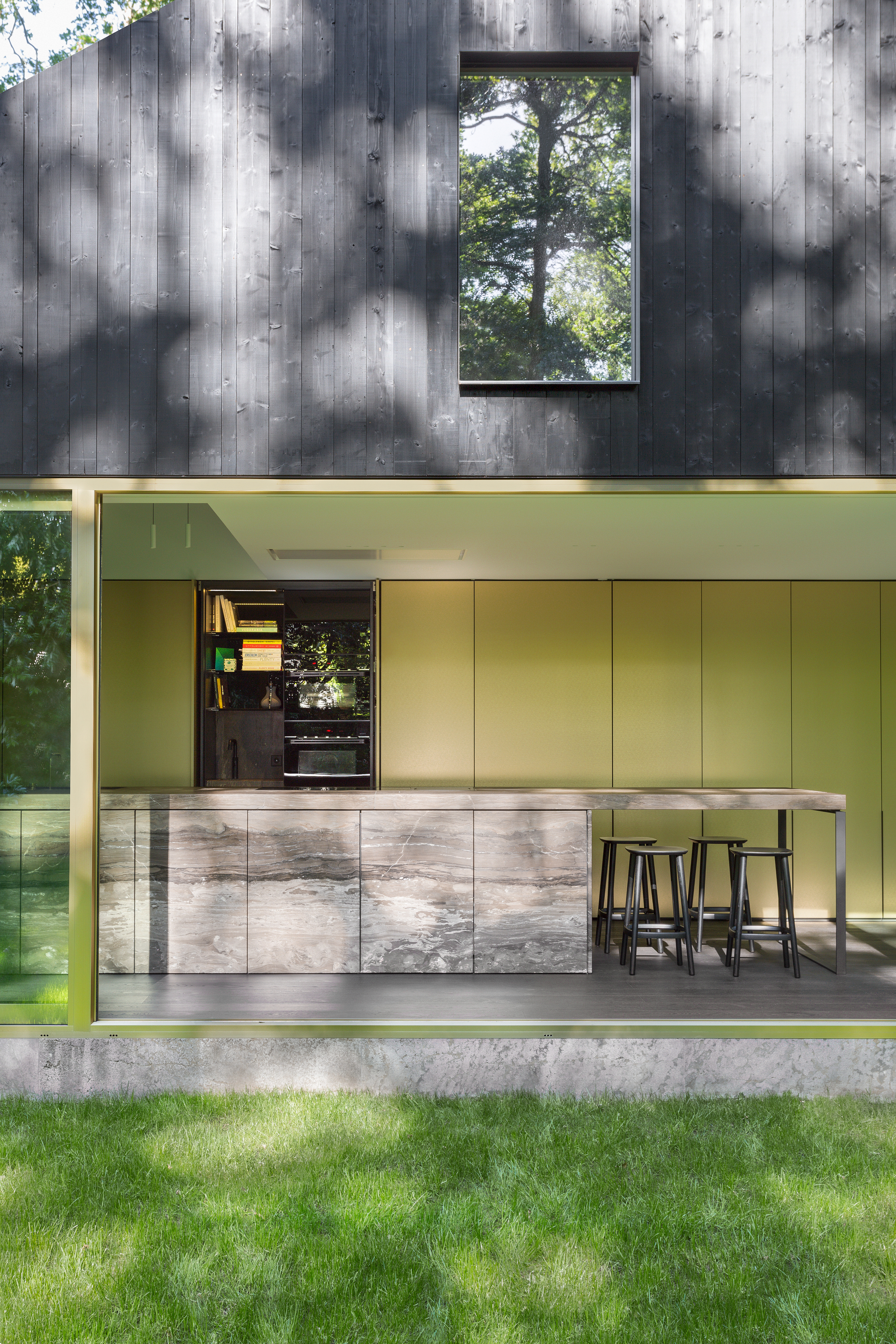 Exterior view showing charred timber upper volume above brass-paneled glazed pavilion and lawn