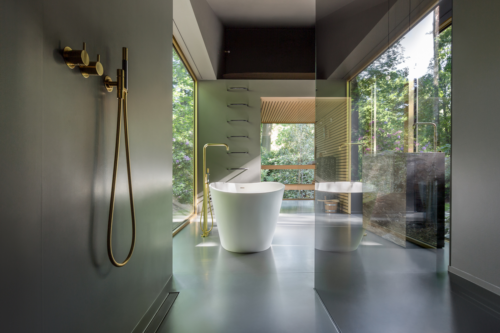 Bathroom with freestanding tub, brass fixtures, and glass walls overlooking the garden at dusk