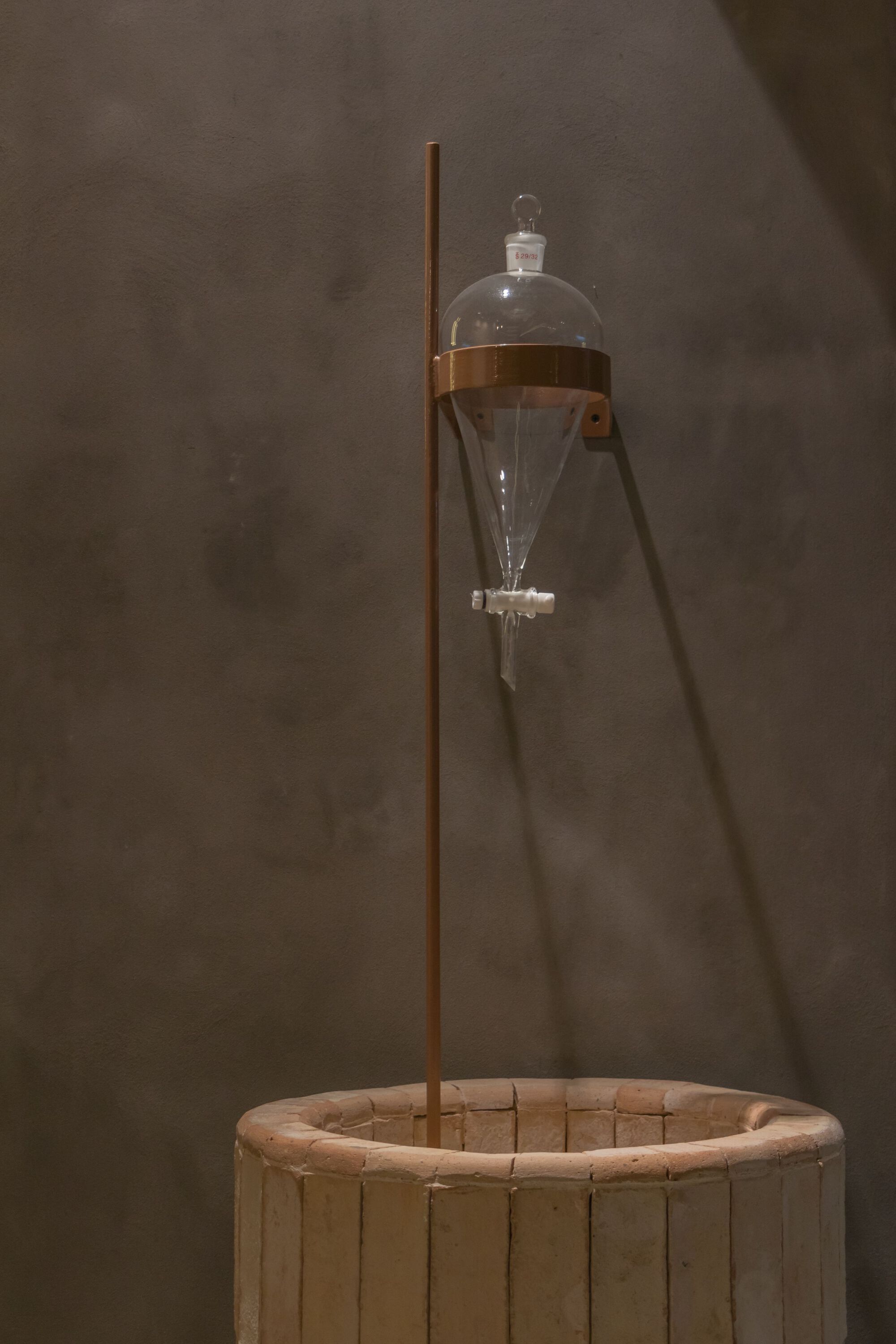Glass separatory funnel suspended over a circular timber basin on a copper stand against plaster wall
