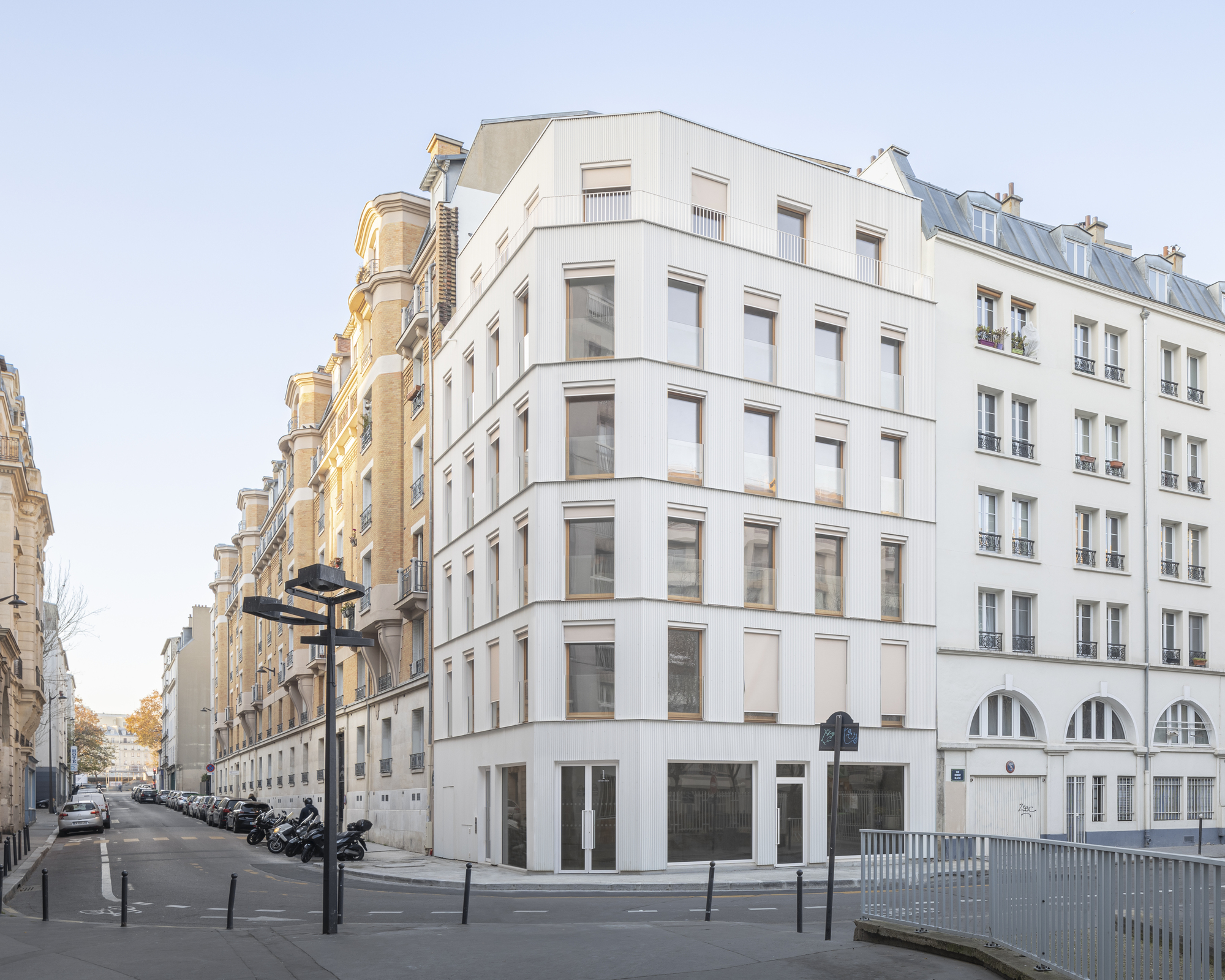 Six-story residential corner building with white rendered facade and arched ground floor openings beside street with parked motorcycles