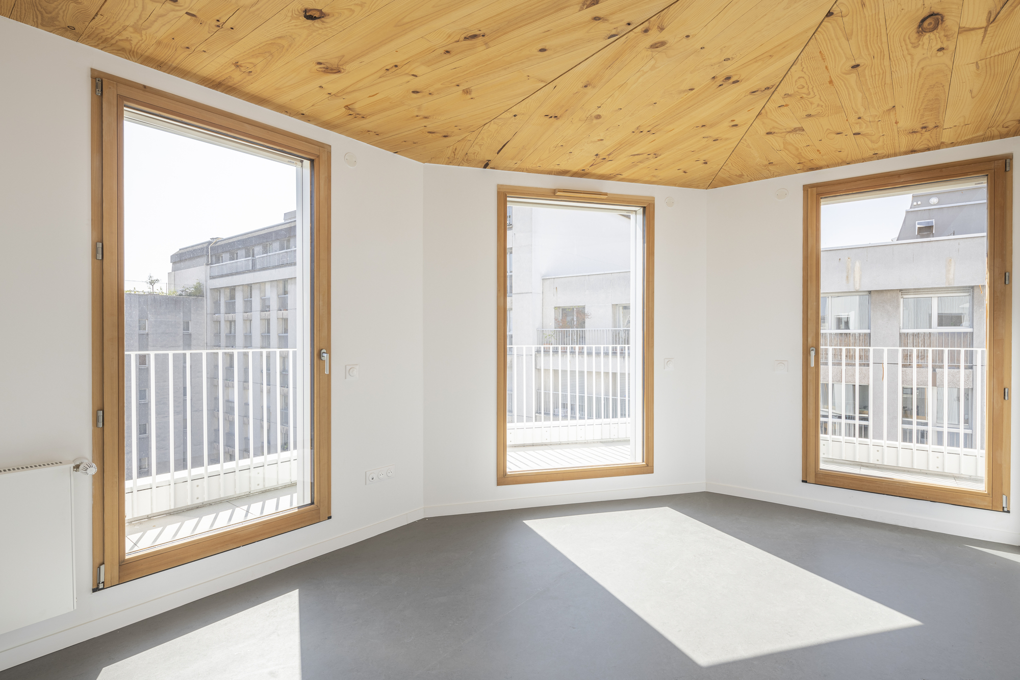 Corner room interior with three timber-framed windows and a plywood plank ceiling in daylight