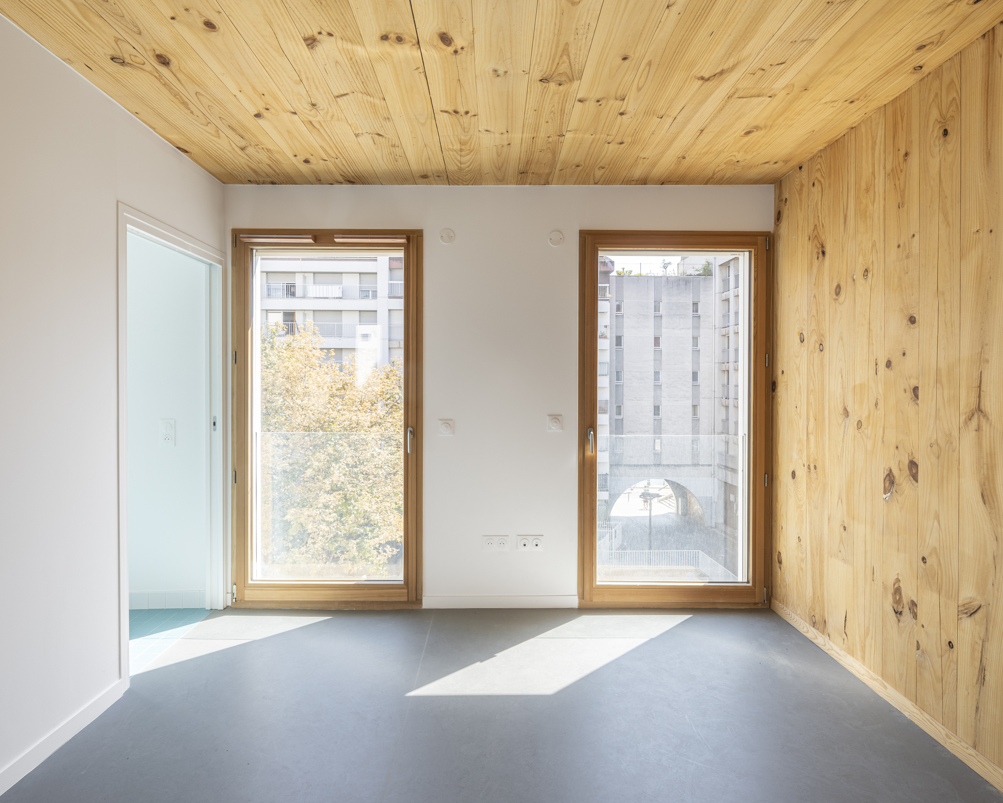 Interior with grey flooring, plywood ceiling and wall, and two windows facing treetops and neighboring buildings