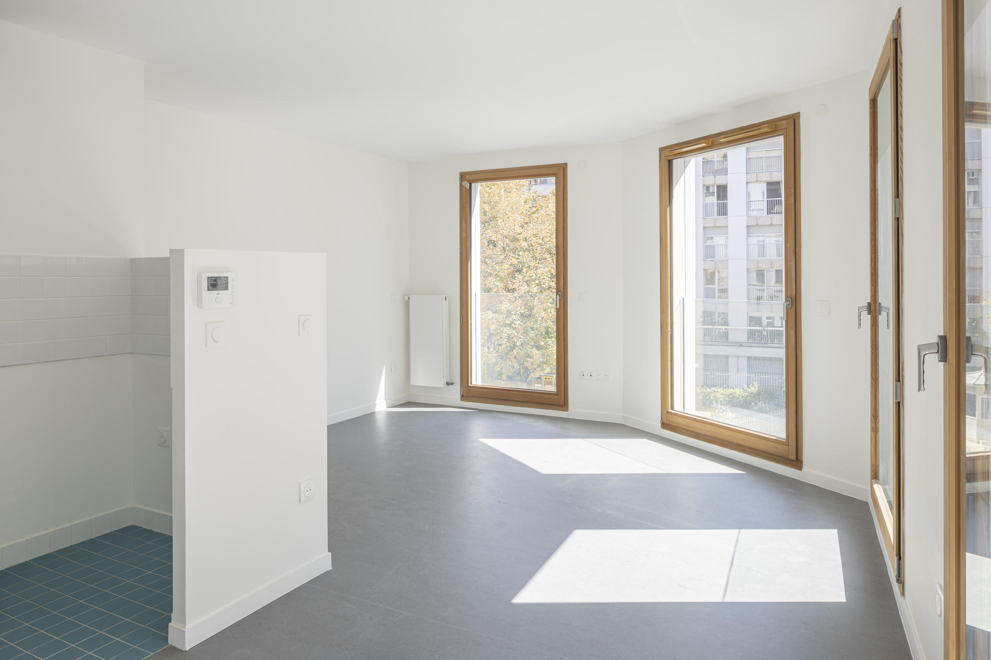 Open plan interior with grey floor, white walls, timber casement windows, and sunlight patches