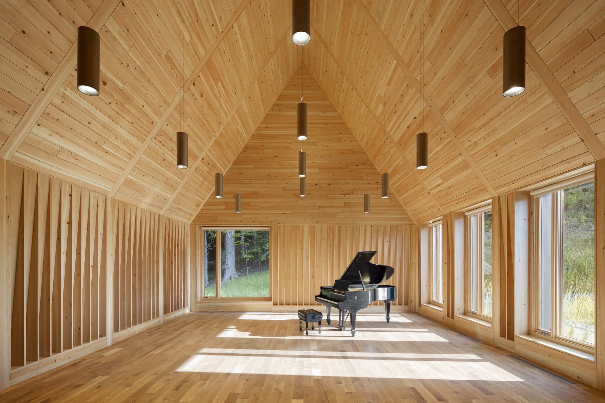 Interior of the timber-clad gable hall with pendant cylinders and a grand piano in sunlight