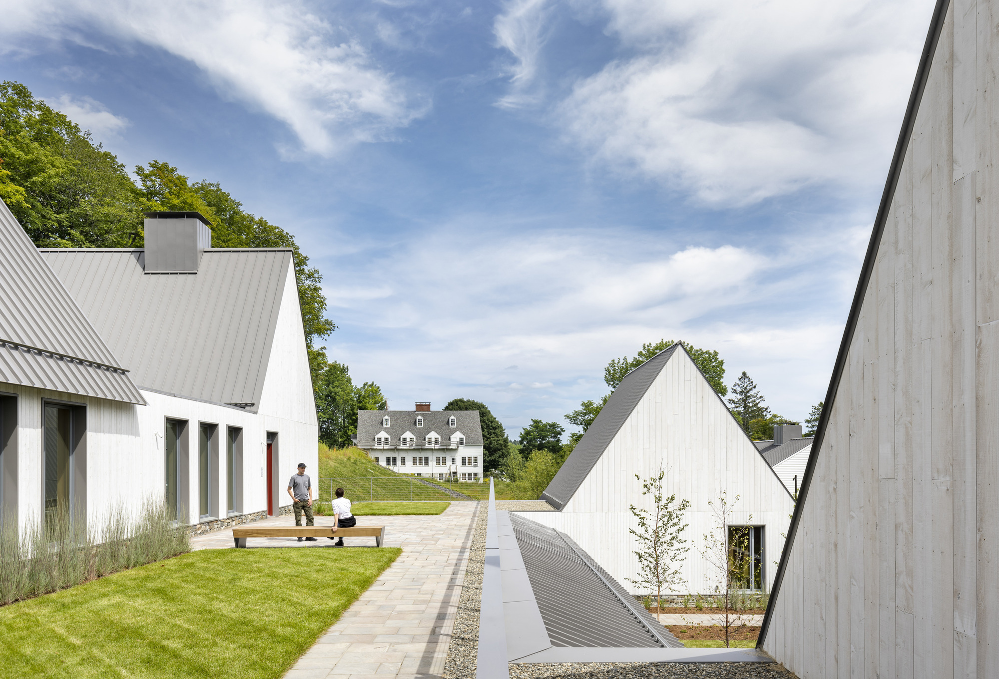 Central paved pathway between gabled volumes with two figures on a lawn under scattered clouds