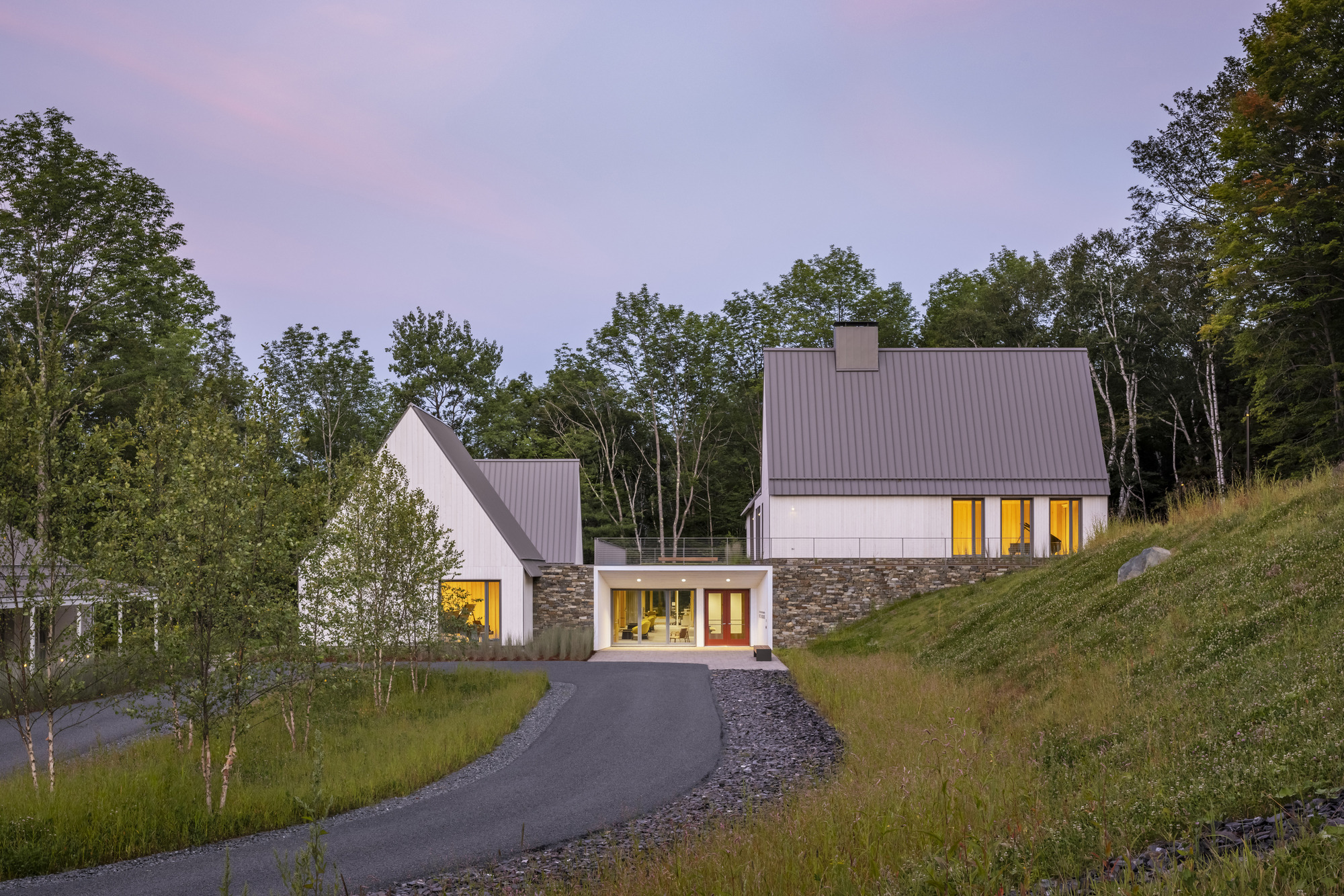Cluster of gabled volumes with stone bases and glowing windows along a sloping drive at twilight