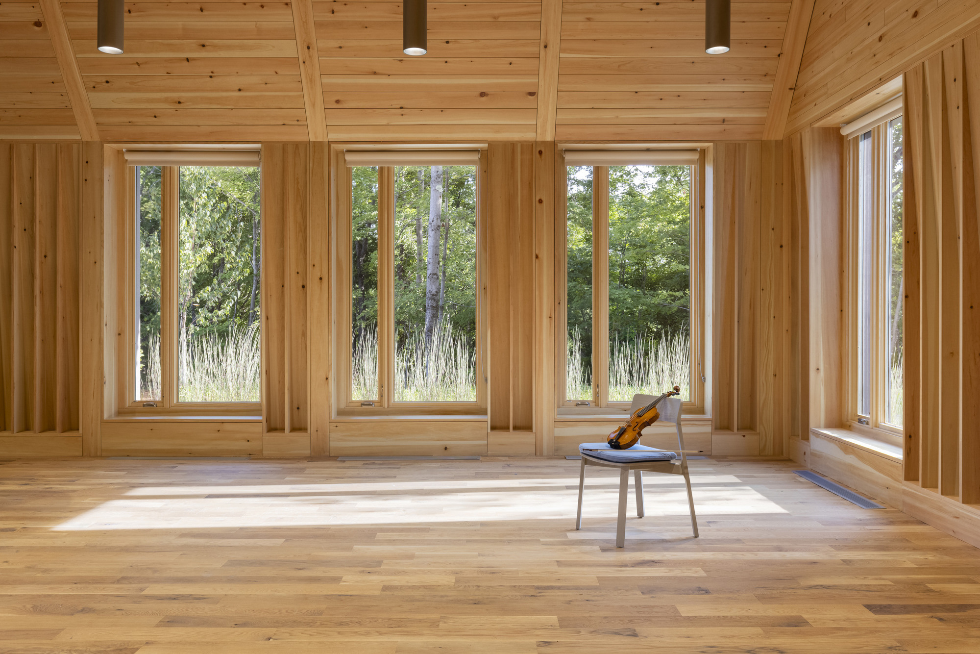 Empty timber-framed room with four tall windows, single chair with violin, and afternoon sunlight across the floor