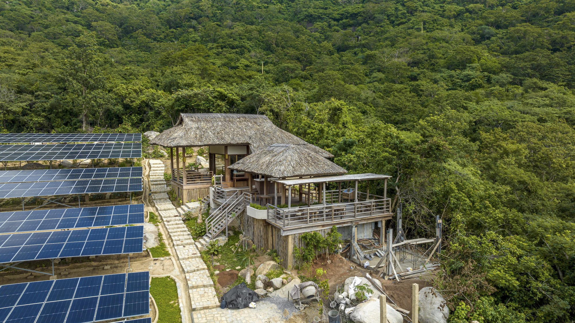 Aerial view of thatched-roof pavilions adjacent to solar panel array amid dense forest