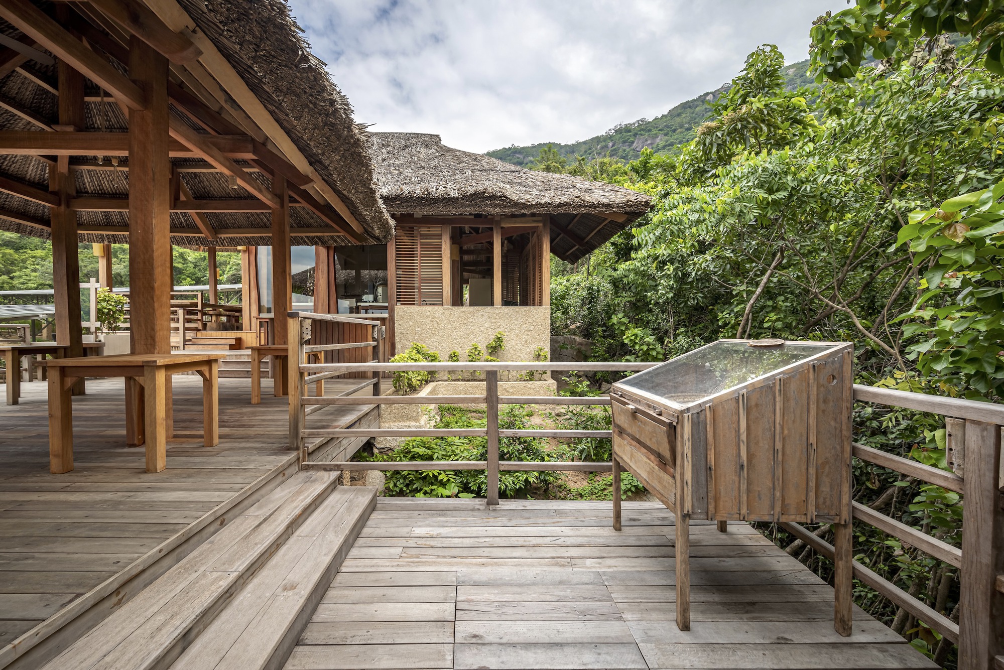 Wooden deck railing and terraced steps leading toward a thatched roof pavilion with green hillside backdrop