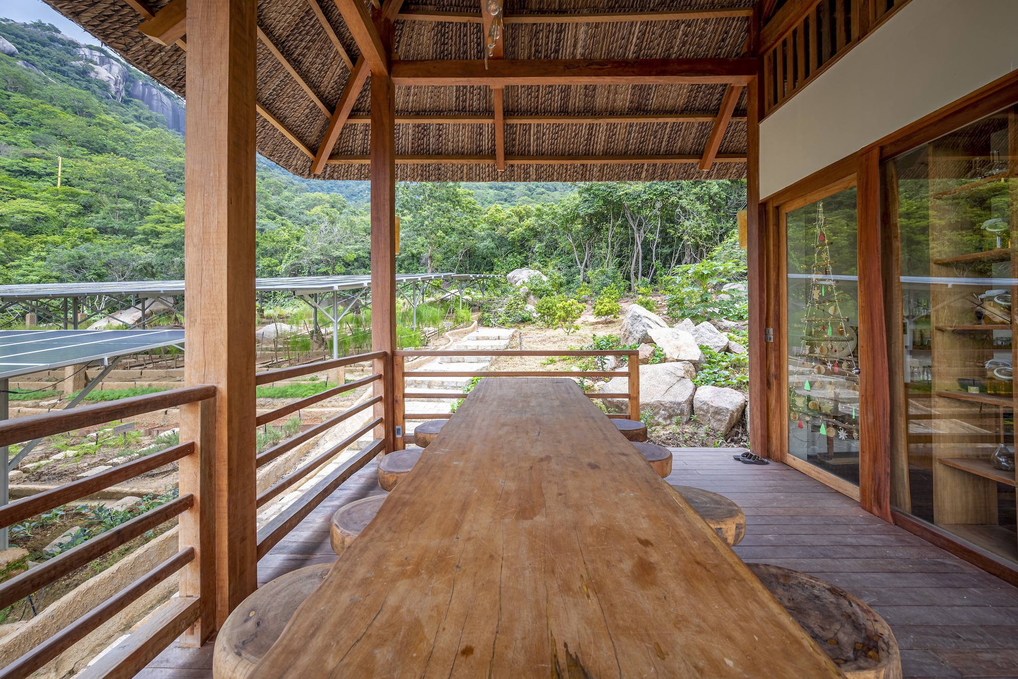 Covered terrace with woven bamboo ceiling and timber columns framing a live-edge table overlooking green hills