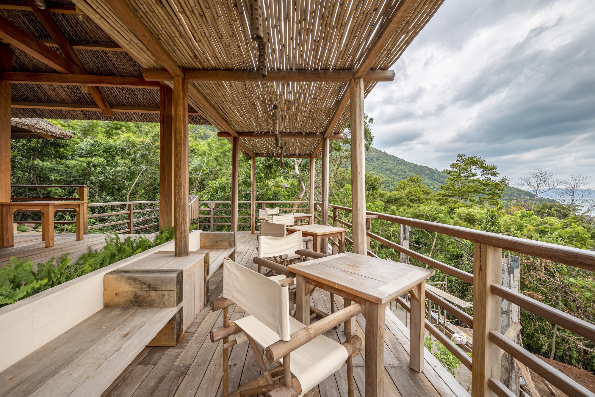 Open-air dining terrace with timber furniture and horizontal rail balustrade under a bamboo-lined roof structure