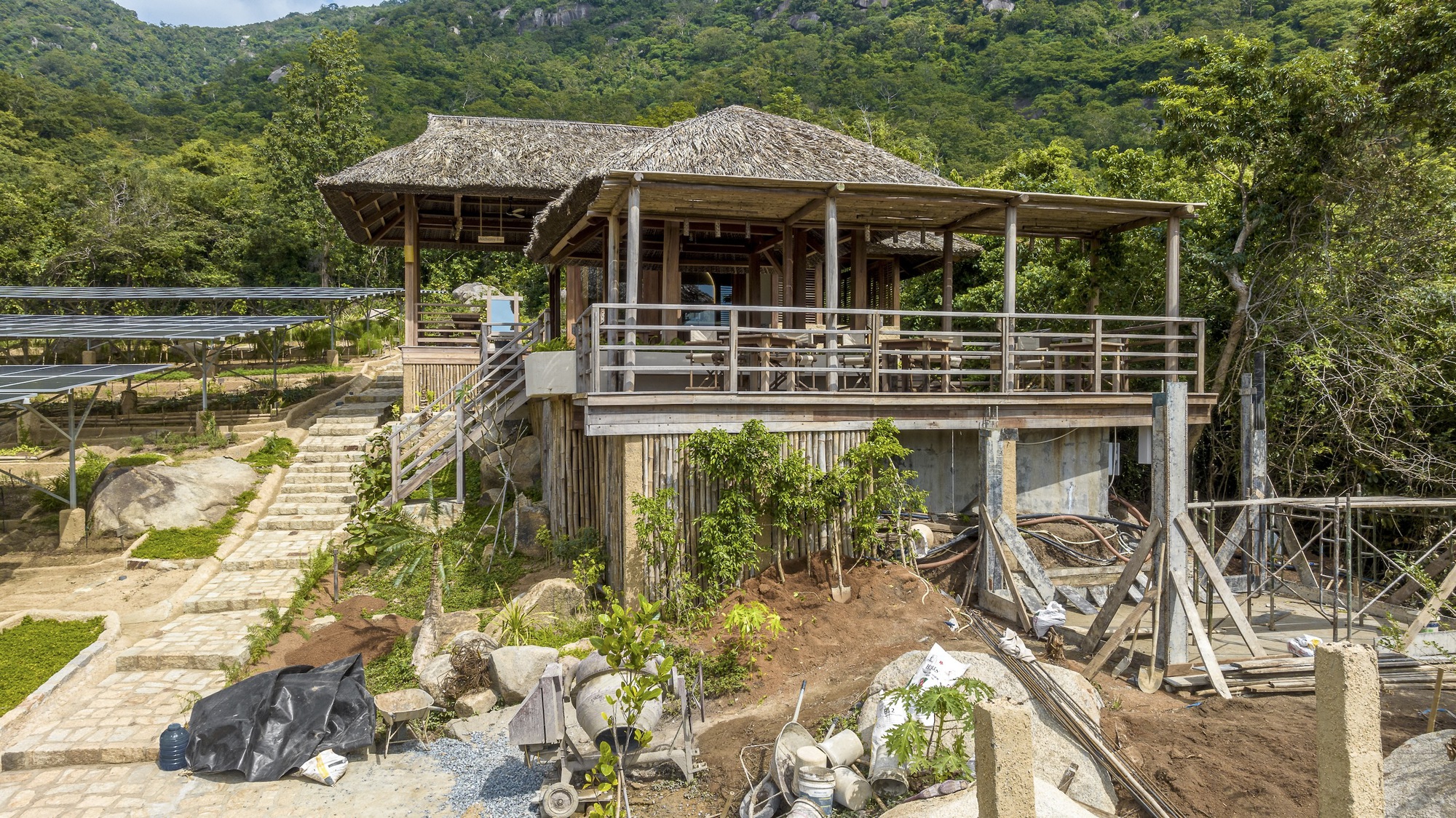Elevated timber pavilion with thatched roofs and horizontal railings above a terraced garden in progress