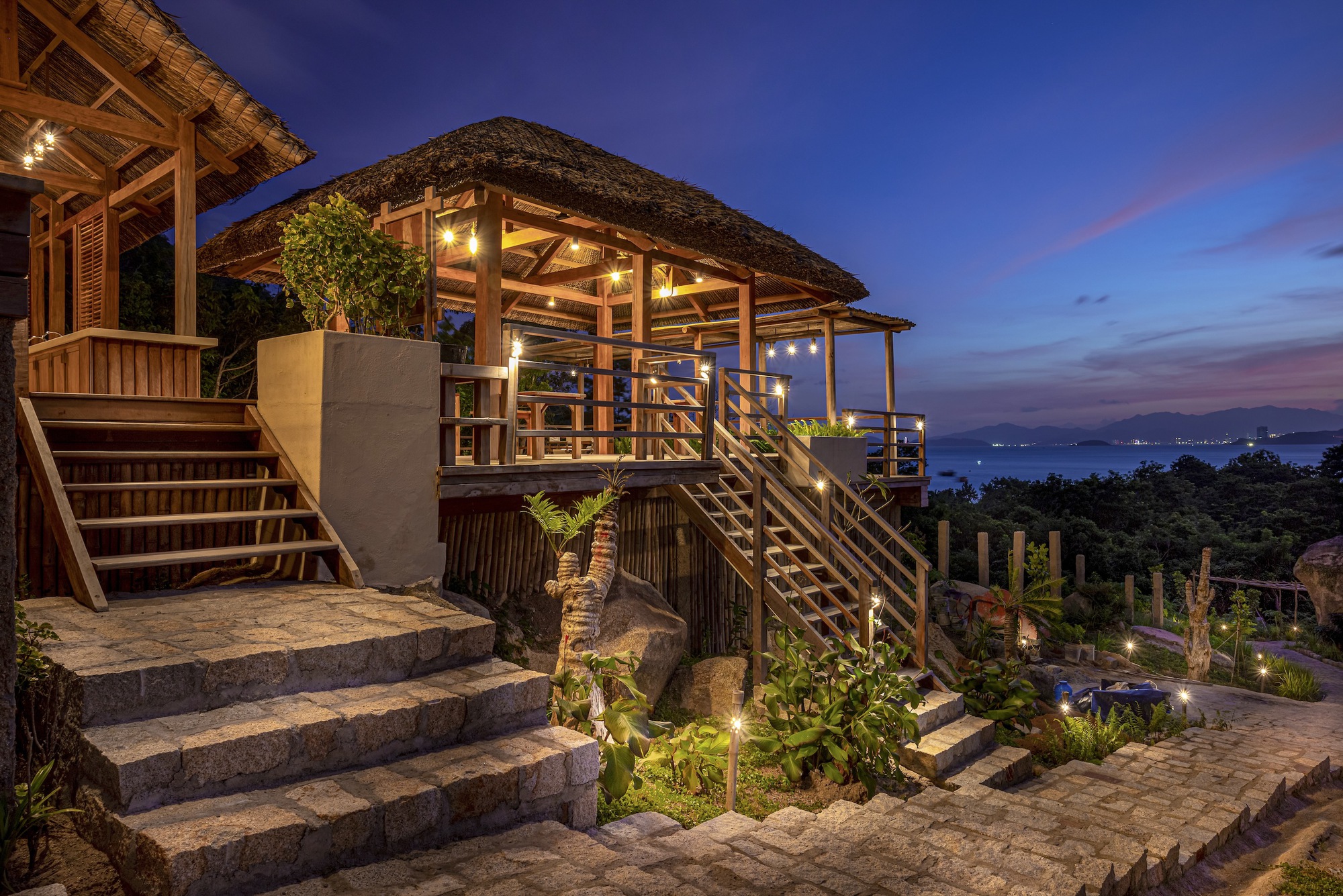 Stone staircase leading to the raised thatched pavilion illuminated at dusk with city lights beyond
