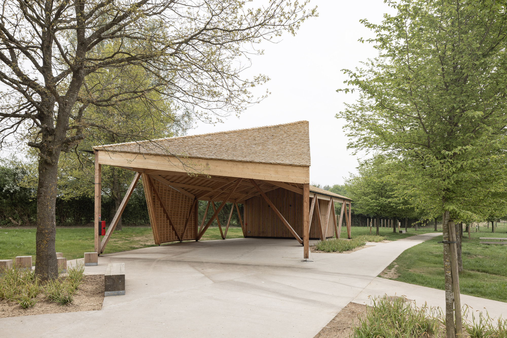 Timber pavilion with diagonal braced columns and woven lattice walls along a concrete pathway in spring