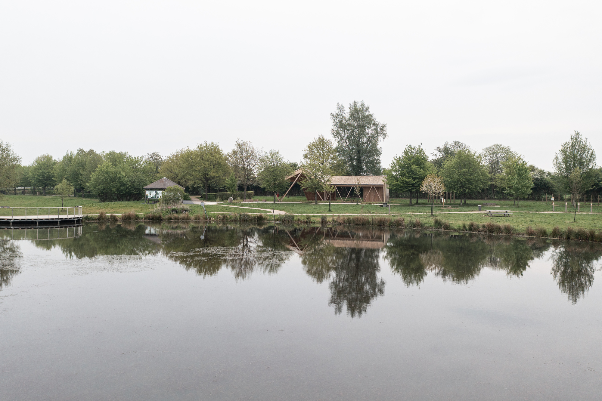 Timber pavilion reflected in the still pond surface with trees lining the grassy shore